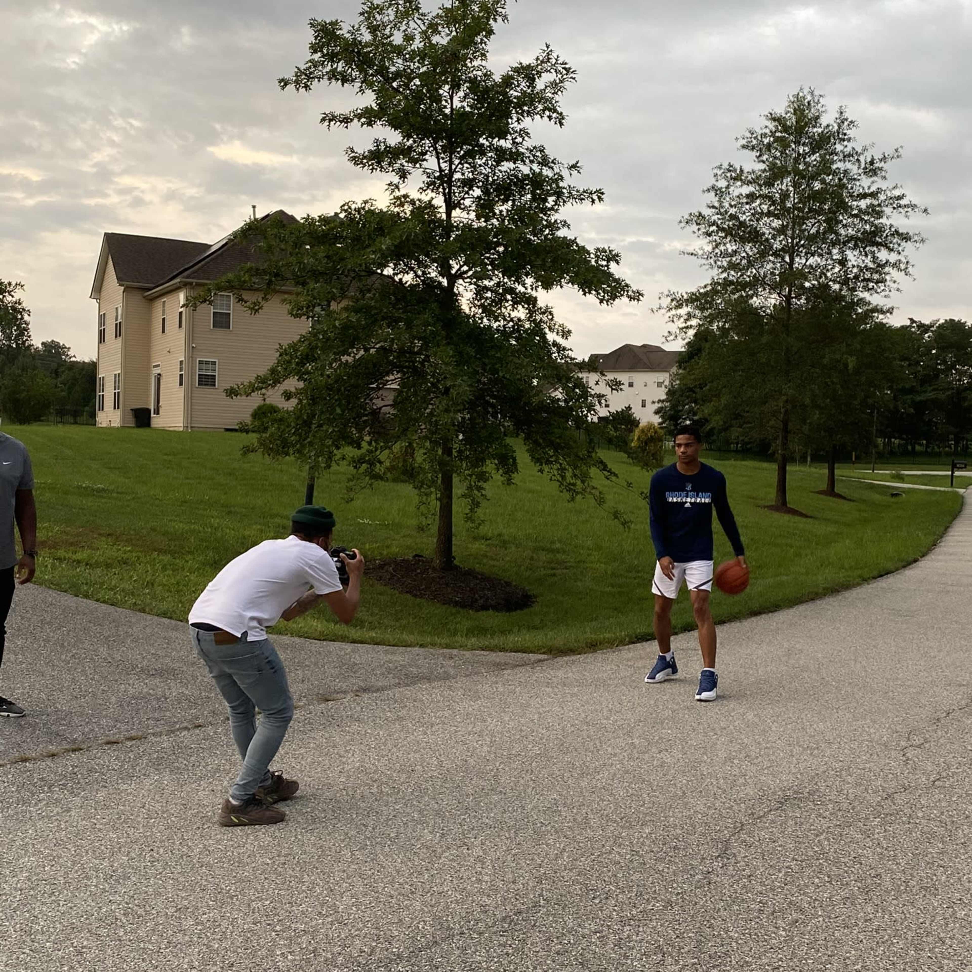 A person holds a basketball while another person takes a photo on a residential street with trees and buildings in the background.