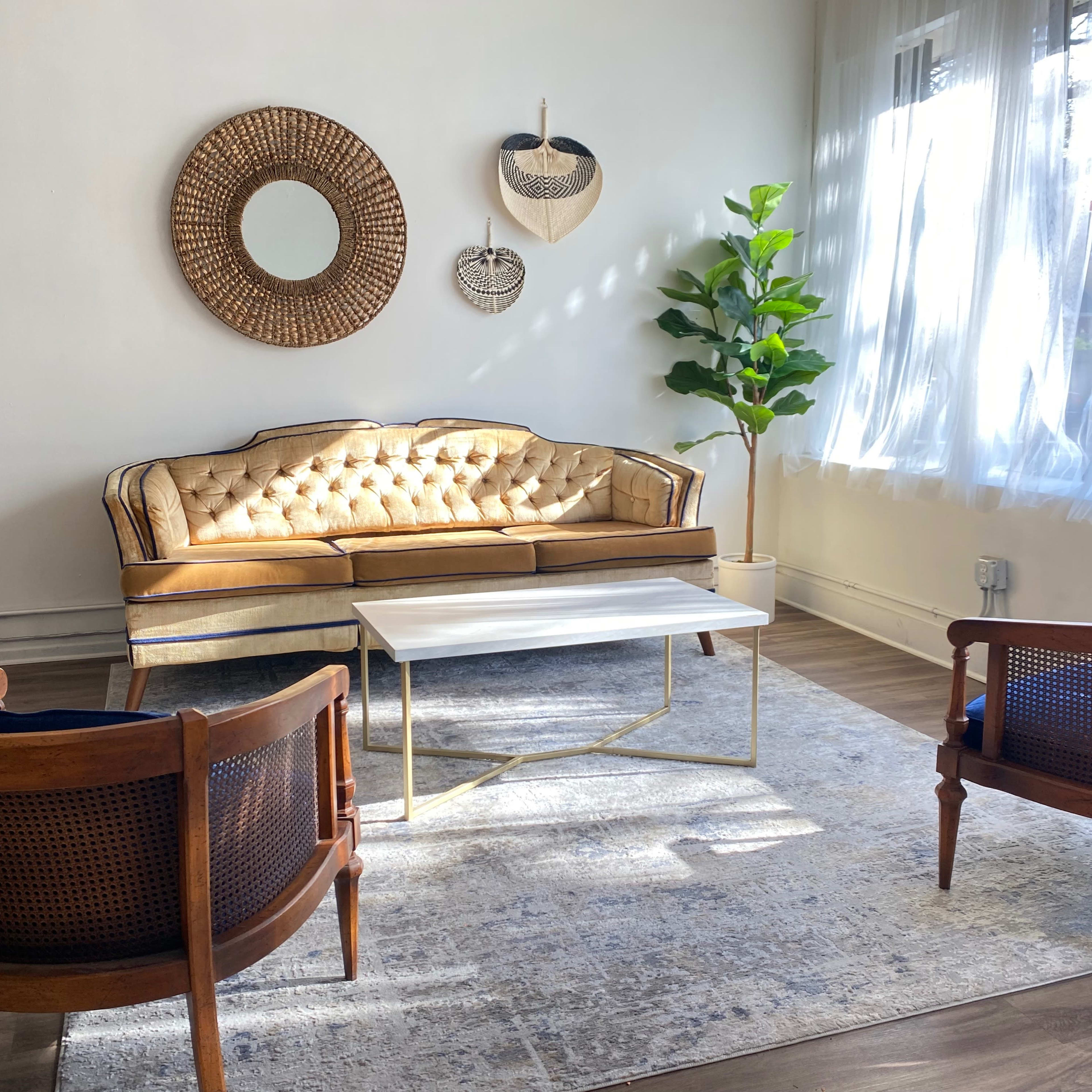 A light-filled living area featuring a tufted sofa, two wooden chairs, a coffee table, and a decorative wall mirror.