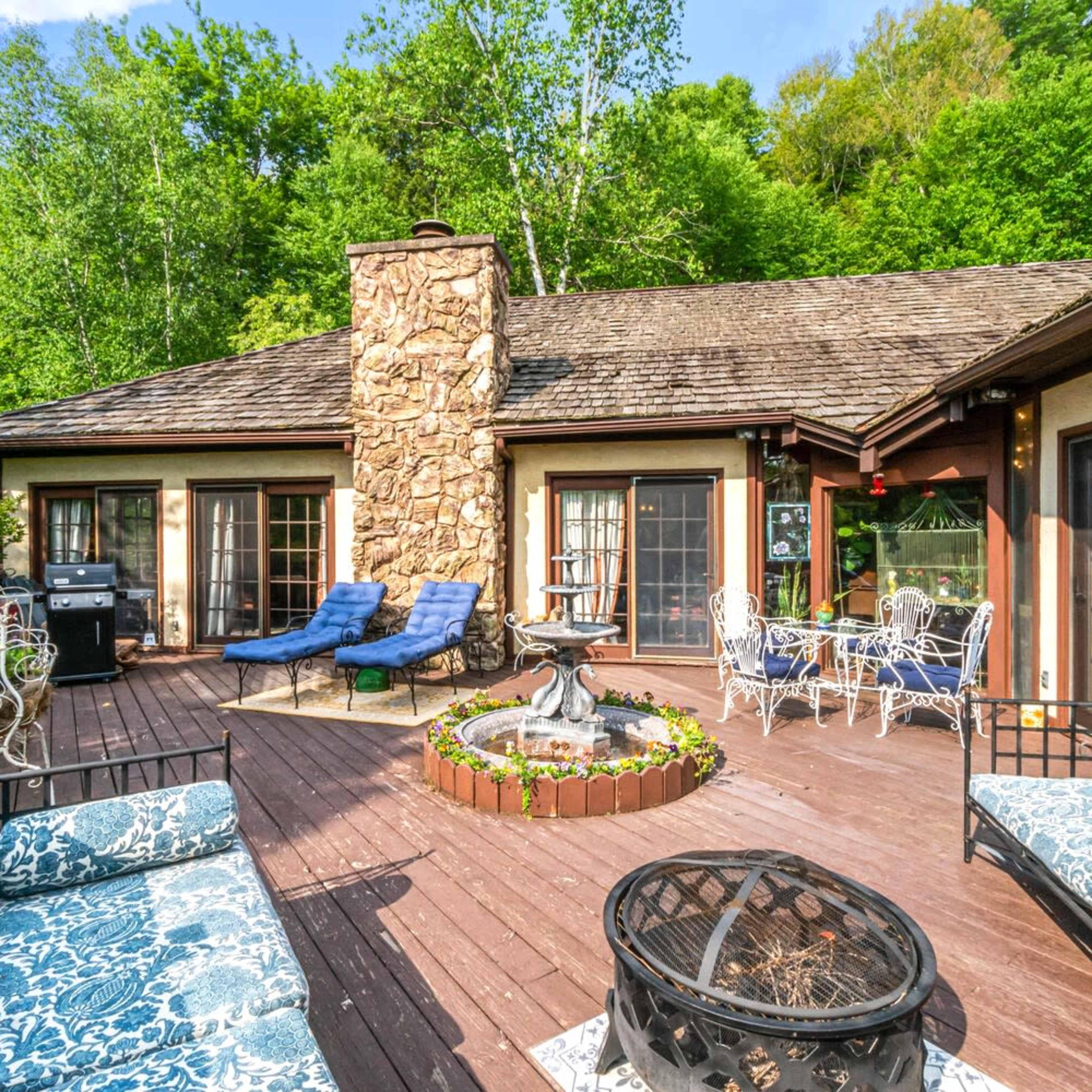 A wooden deck with seating areas, a fire pit, and a stone chimney, surrounded by lush greenery.