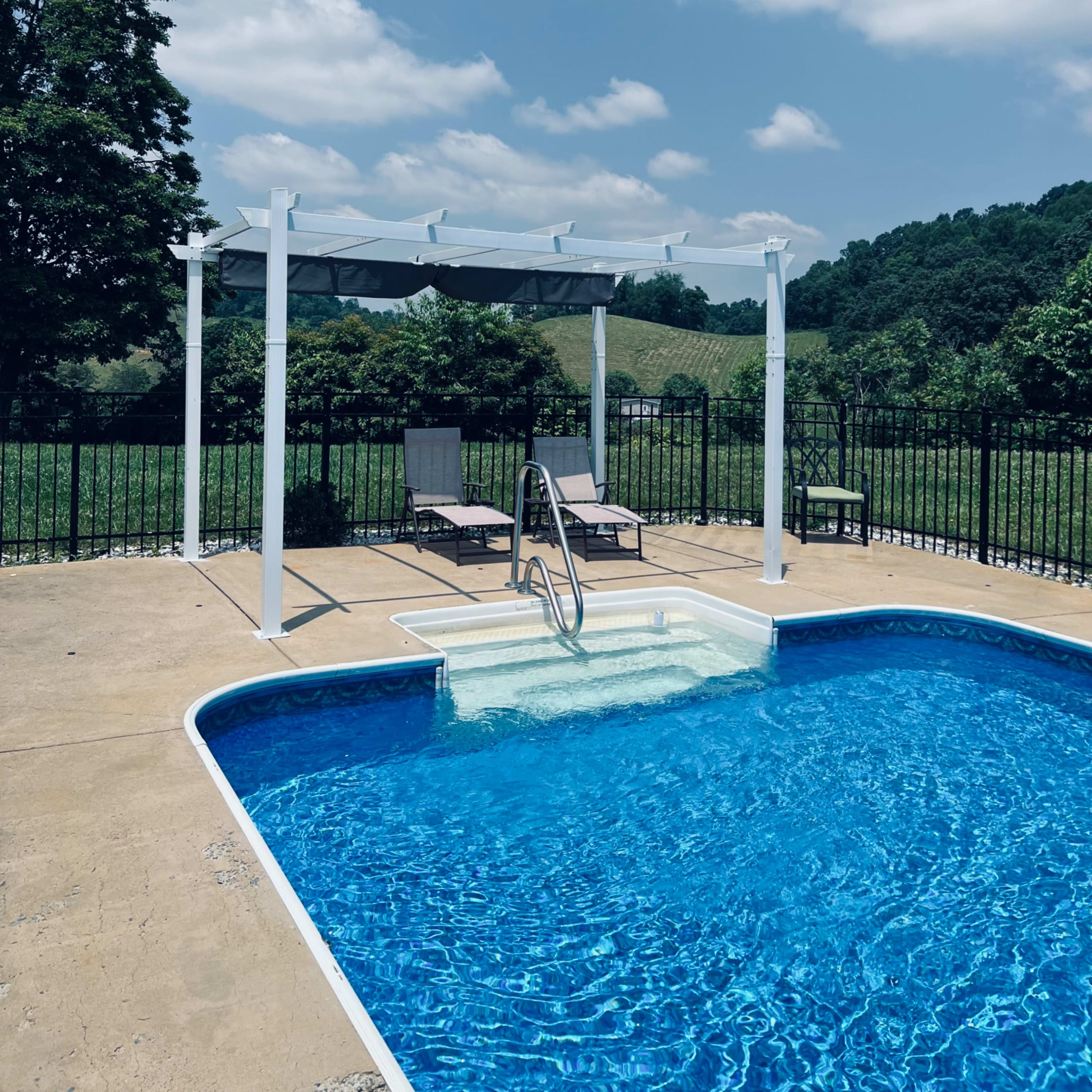 A swimming pool with a ladder and lounge chairs is situated beside a pergola, surrounded by a fenced backyard and green hills under a partly cloudy sky.