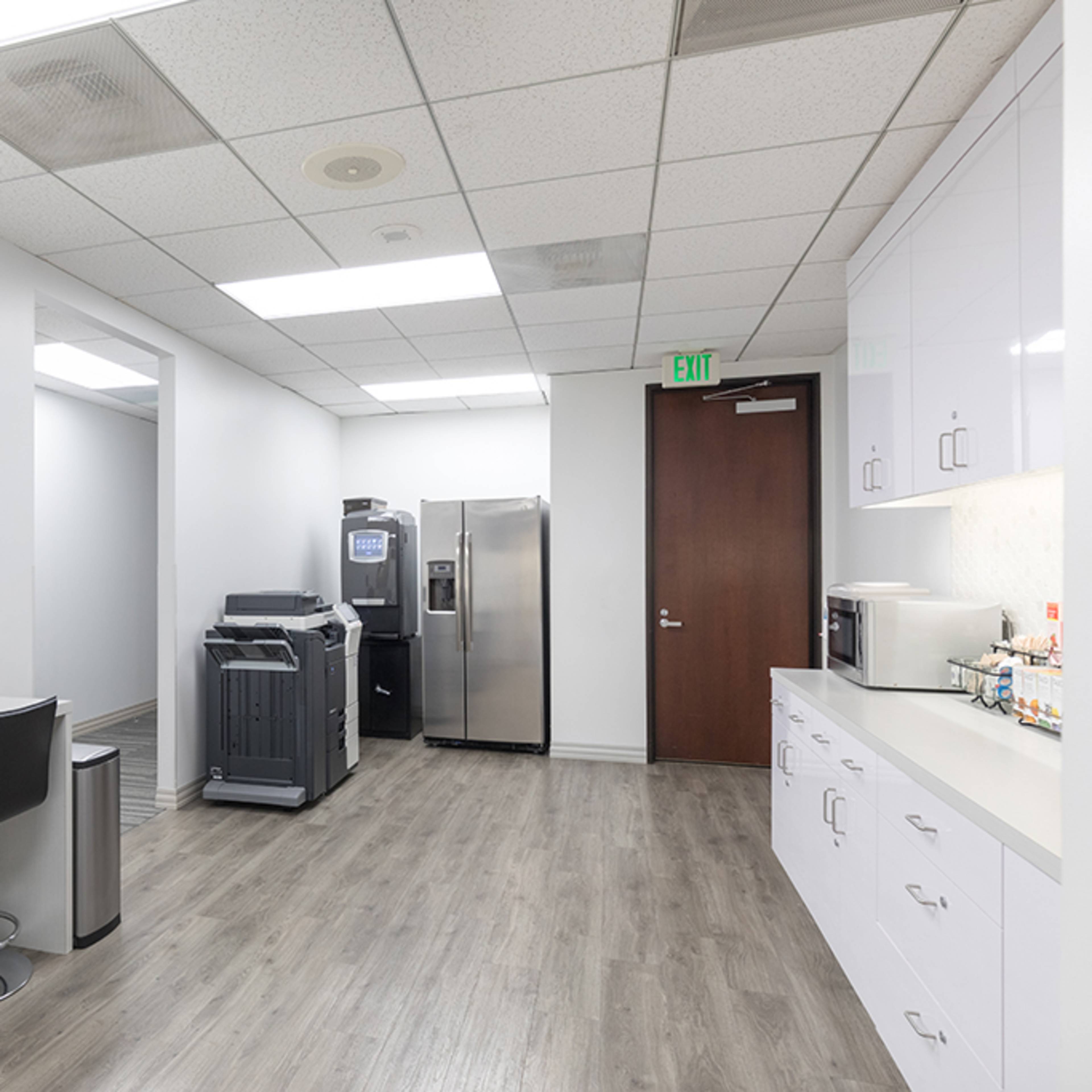 A modern break room with a seating area, refrigerator, microwave, and a printer, featuring light-colored walls and a colorful abstract painting.