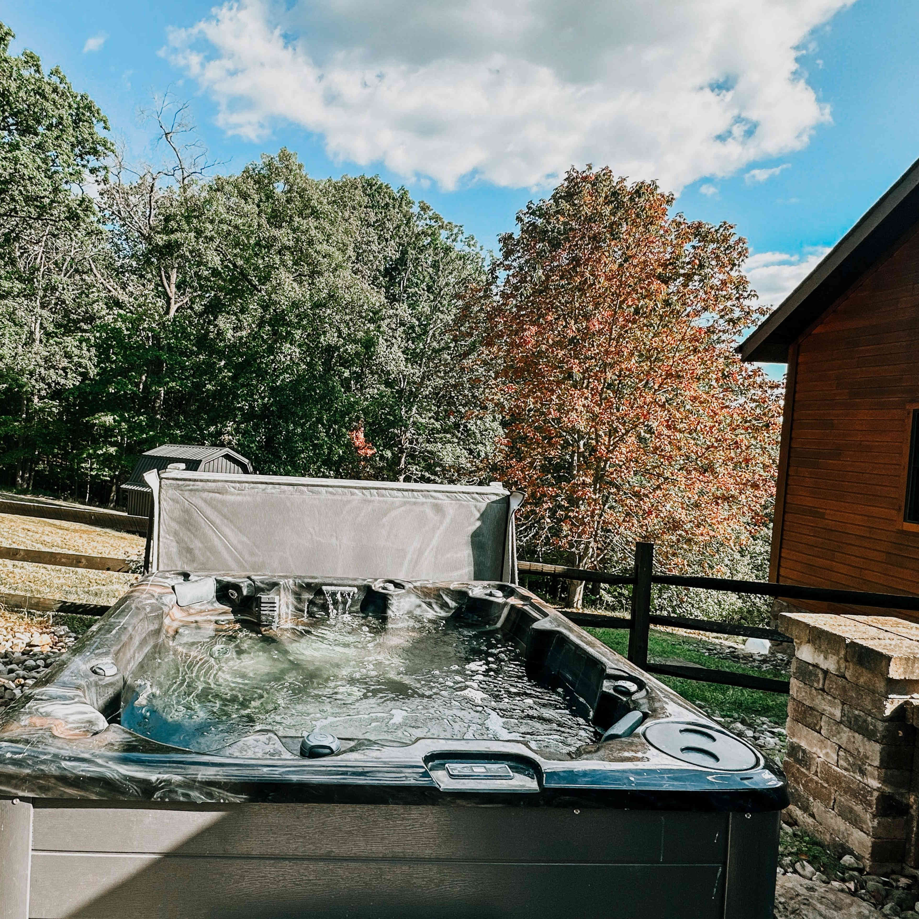 A hot tub filled with water is situated next to a wooden fence and surrounded by trees with autumn foliage.
