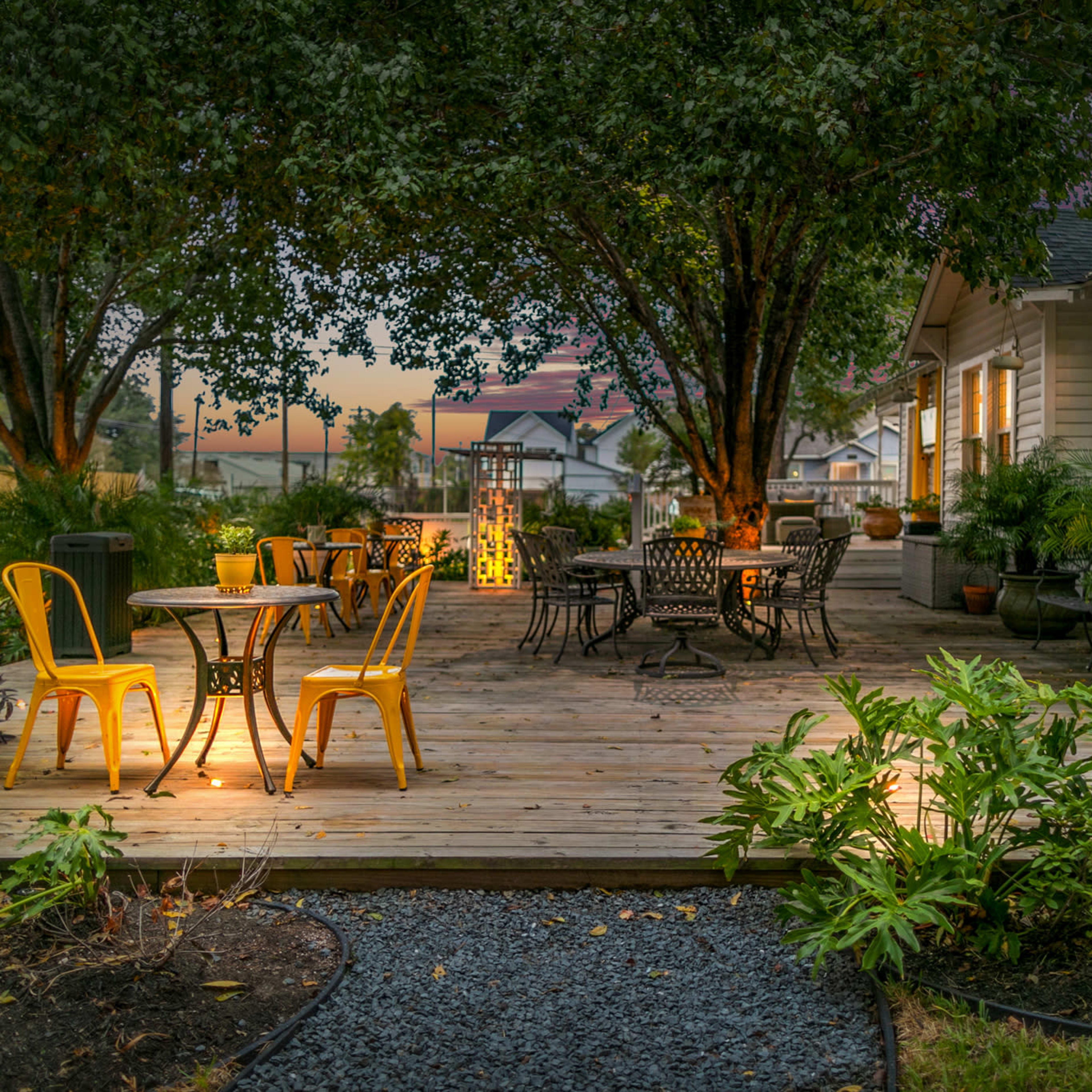 A wooden deck with several tables and chairs surrounded by plants and trees, illuminated by warm lighting from the windows of a nearby house.