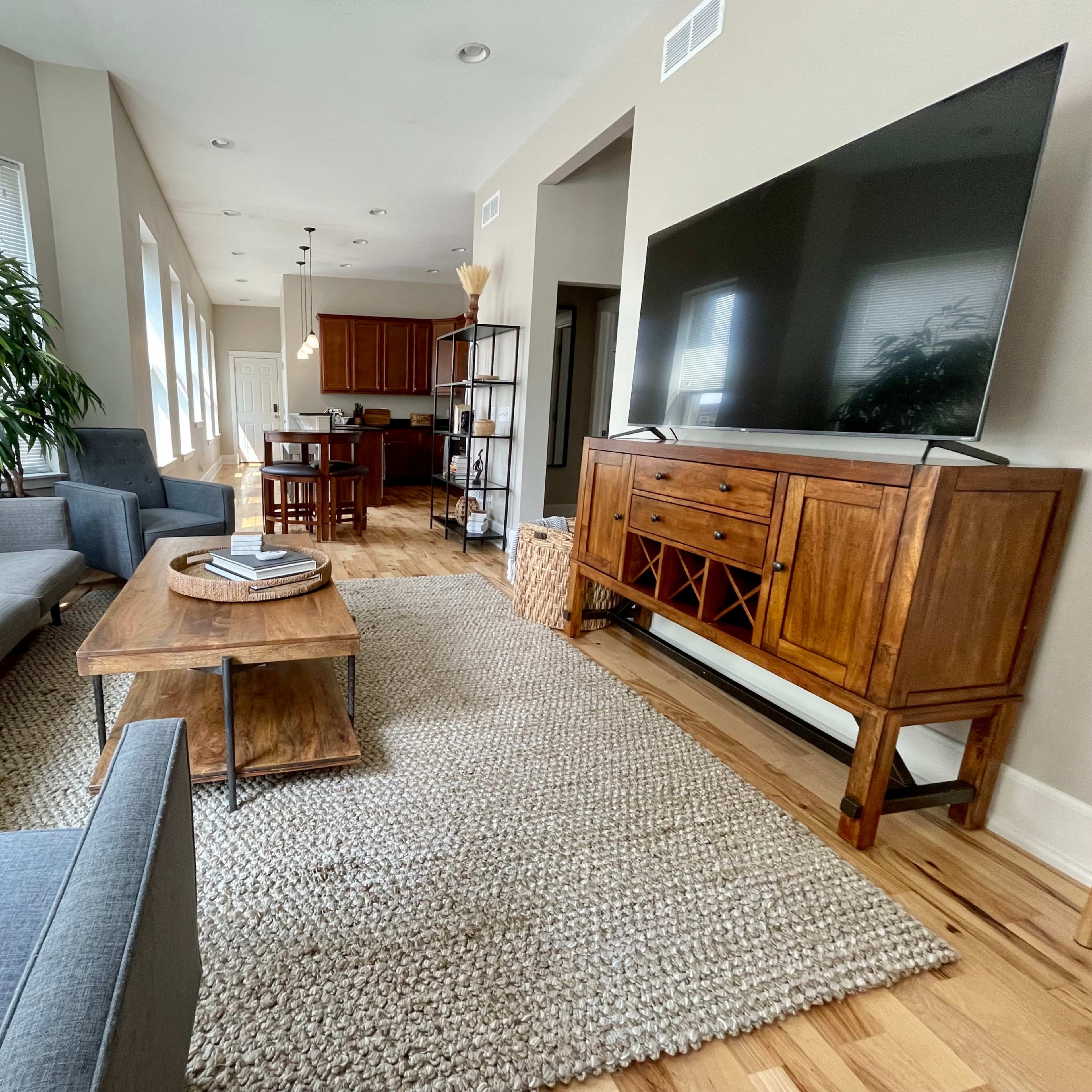 A modern living room features a gray sofa, a wooden coffee table, a large television, and a dining area visible in the background.