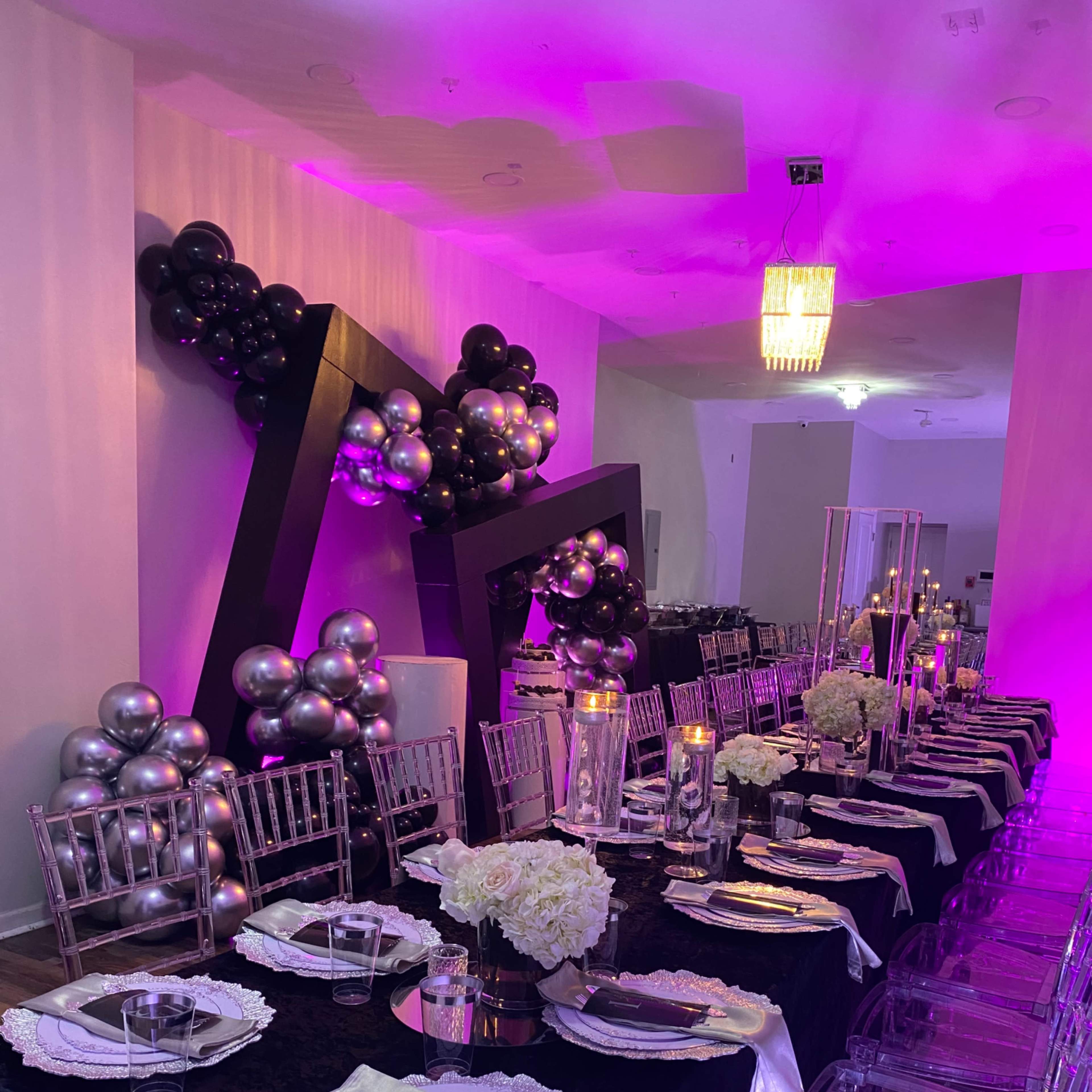 A long dining table is set in a venue adorned with black, silver, and purple decorations, featuring clear acrylic chairs and floral centerpieces.