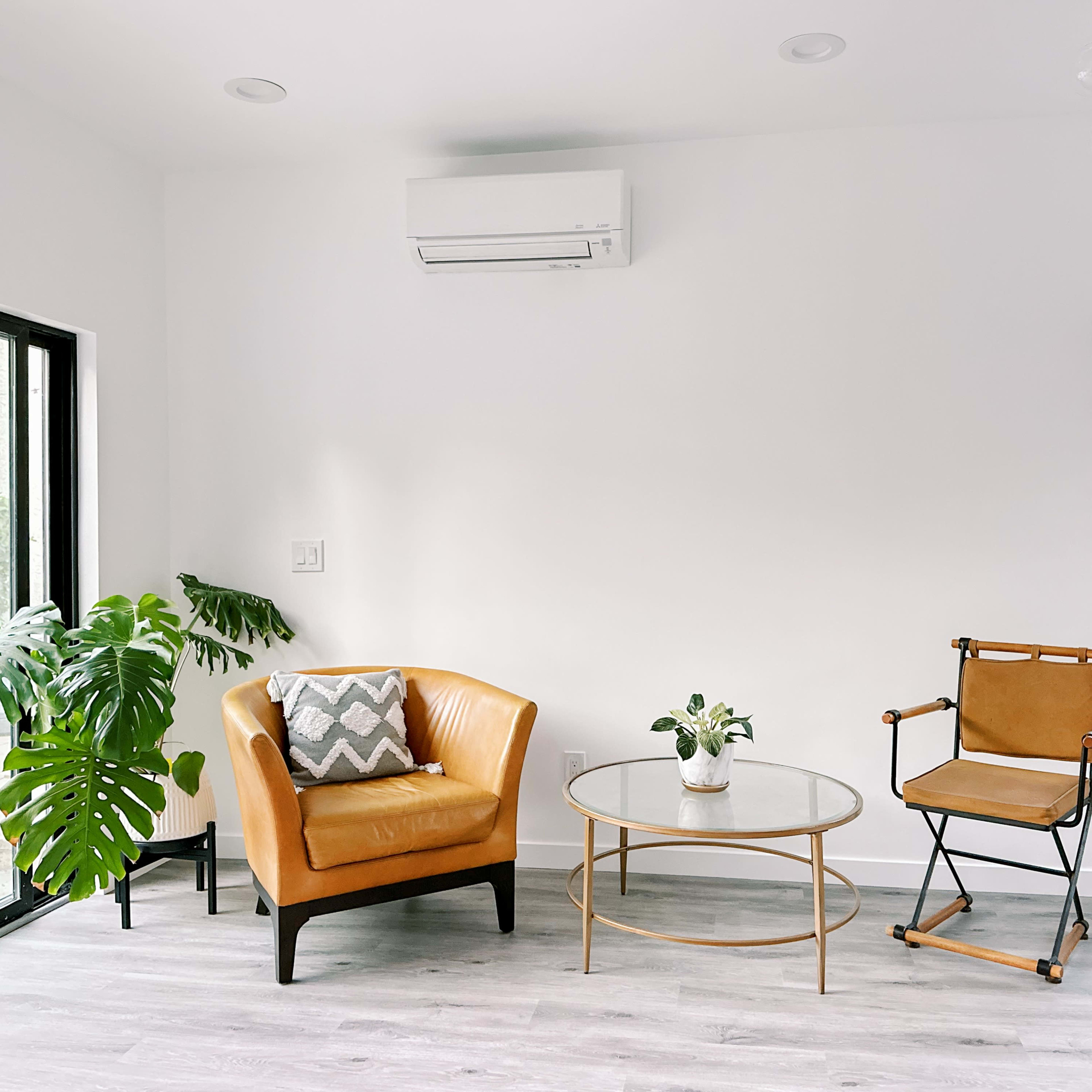 The scene features a bright, modern living area with a brown armchair, a glass coffee table, and a folding chair, surrounded by greenery and large windows.