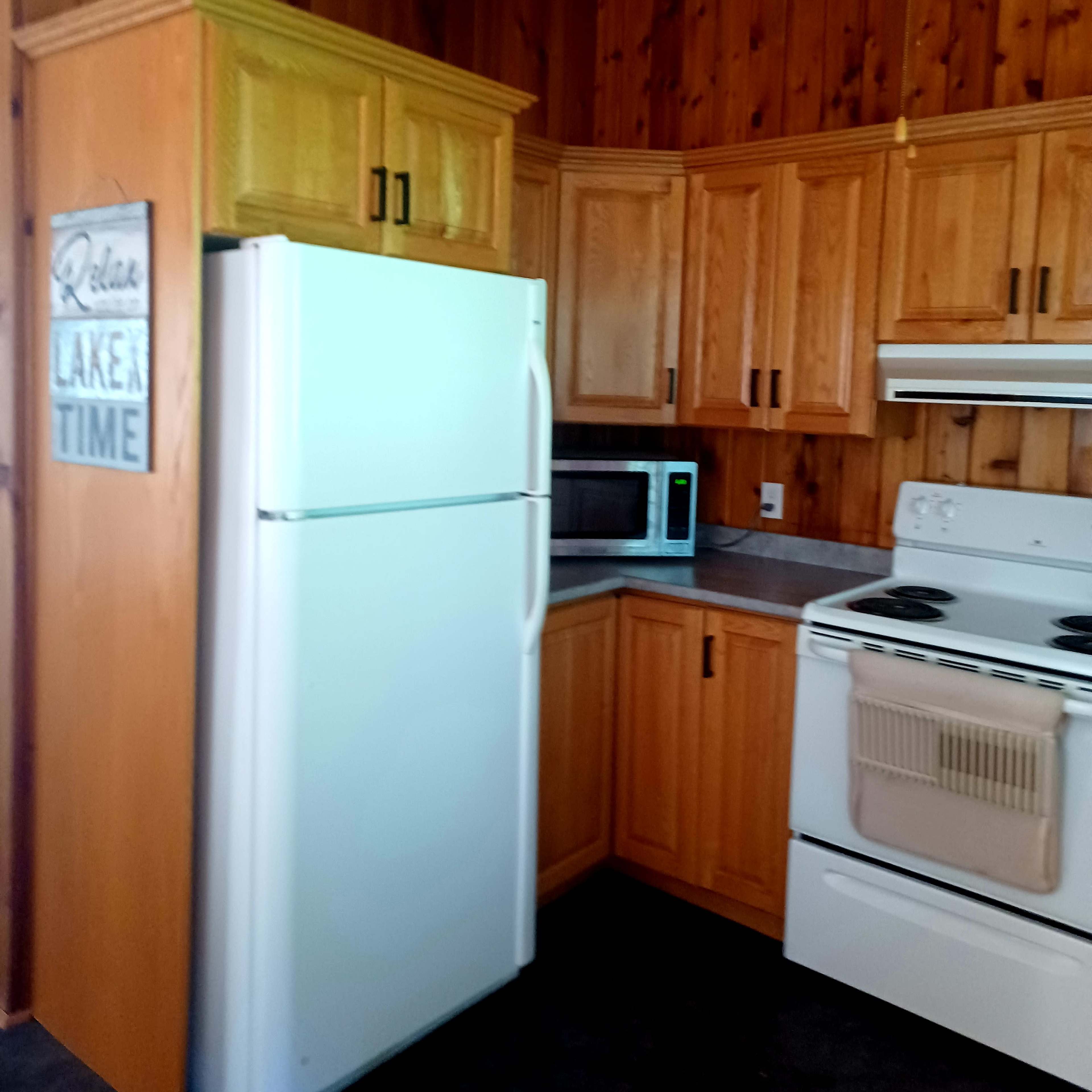 A kitchen with wooden cabinetry, a white refrigerator, a microwave, and a stove against a paneled wall.