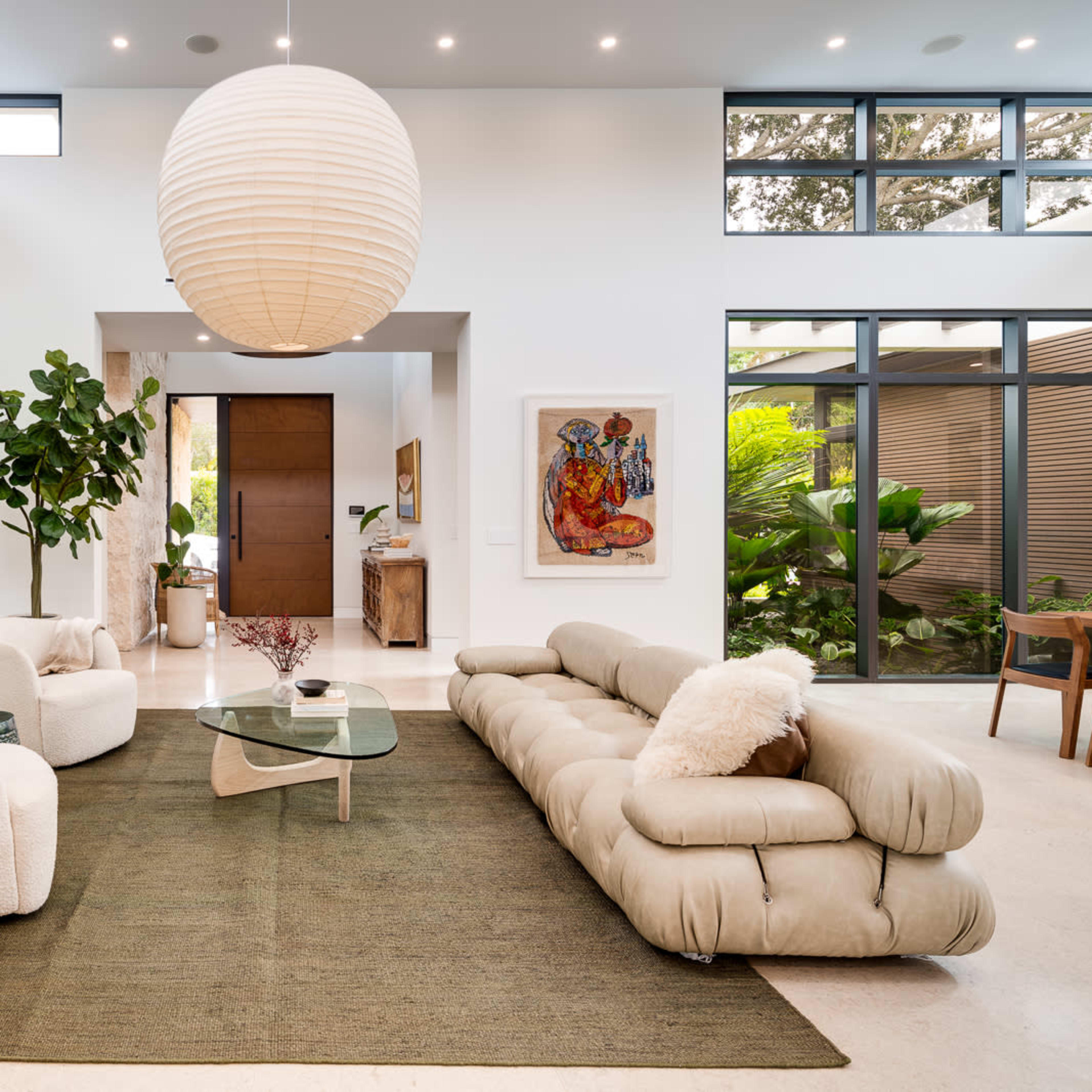 The living room features a contemporary sofa, two plush armchairs, a glass coffee table, and large windows allowing natural light to illuminate the space.