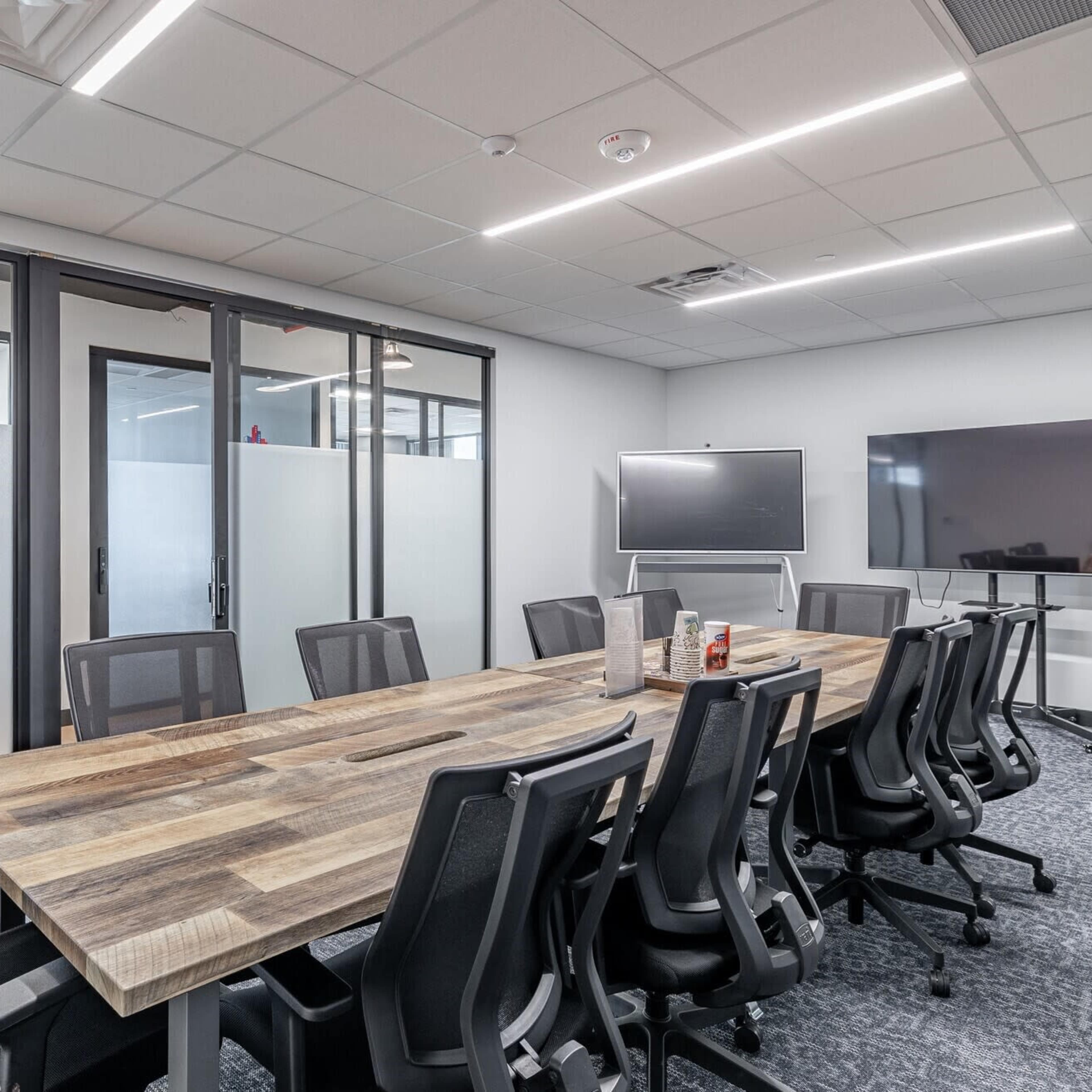 A modern conference room features a long wooden table surrounded by black office chairs, with glass partitions and large screens on the walls.