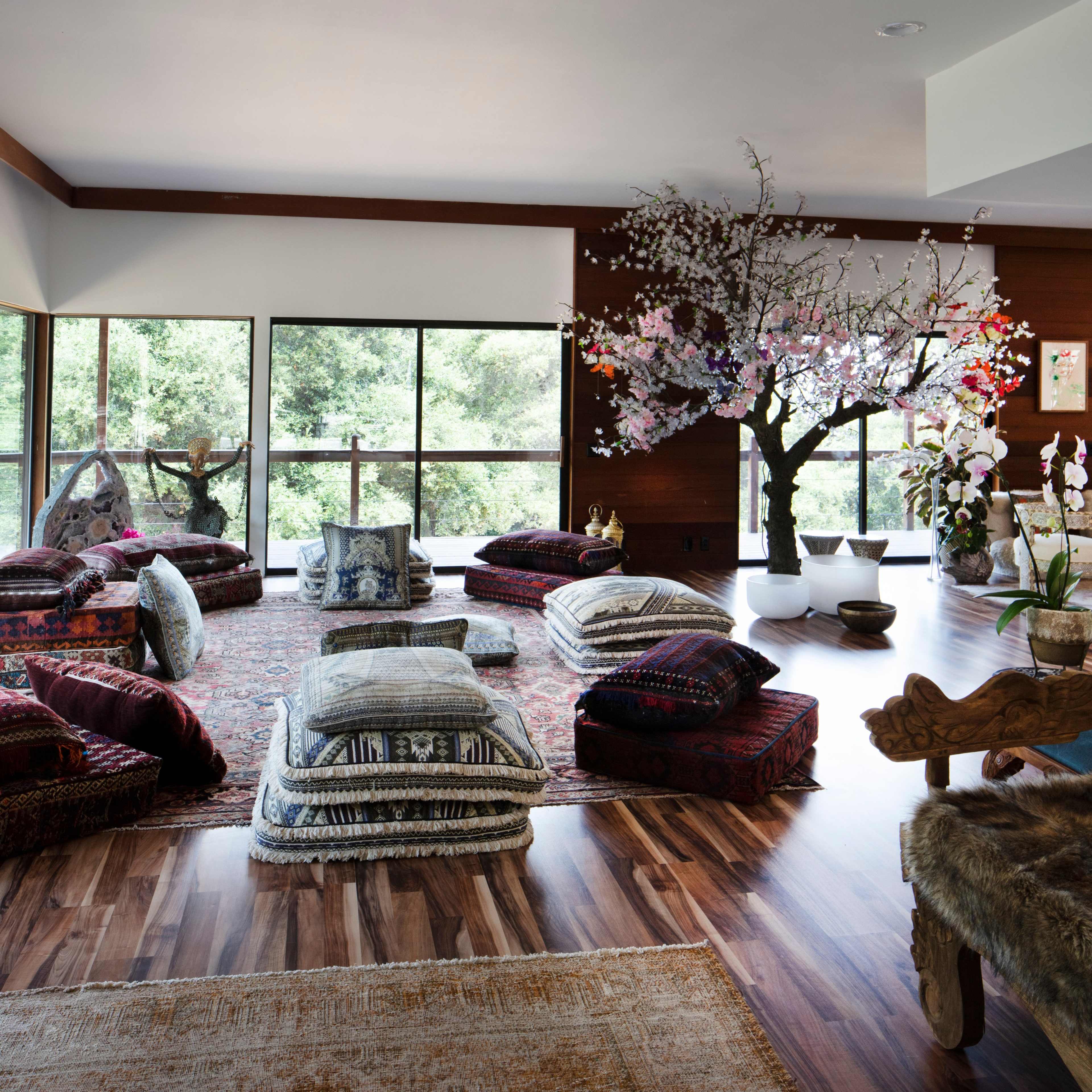 A spacious room is filled with colorful cushions arranged on a patterned rug, with large windows offering views of greenery and a decorative tree in one corner.