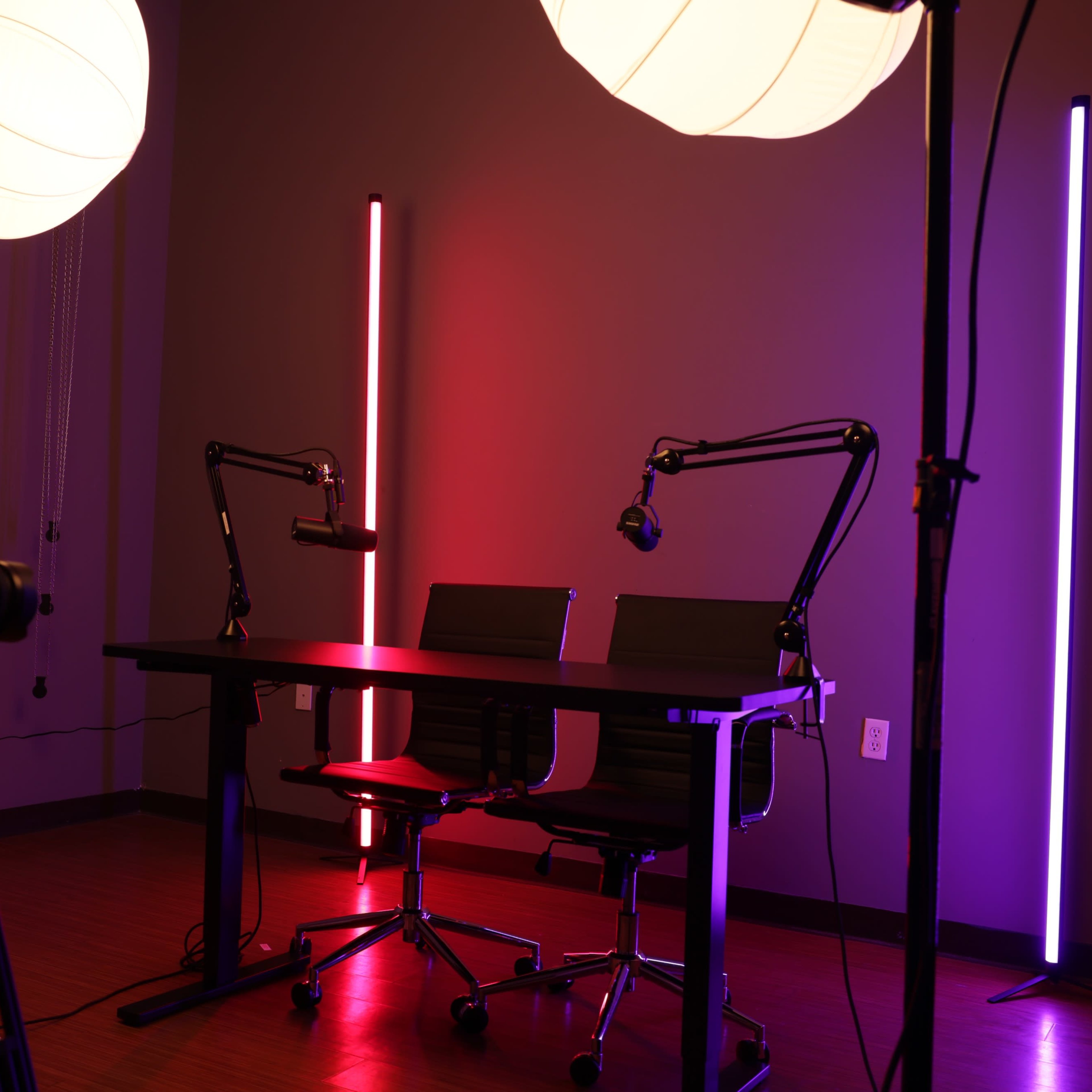 A modern studio setup features a table with two chairs, illuminated by colorful neon lights and equipped with microphones on adjustable arms.
