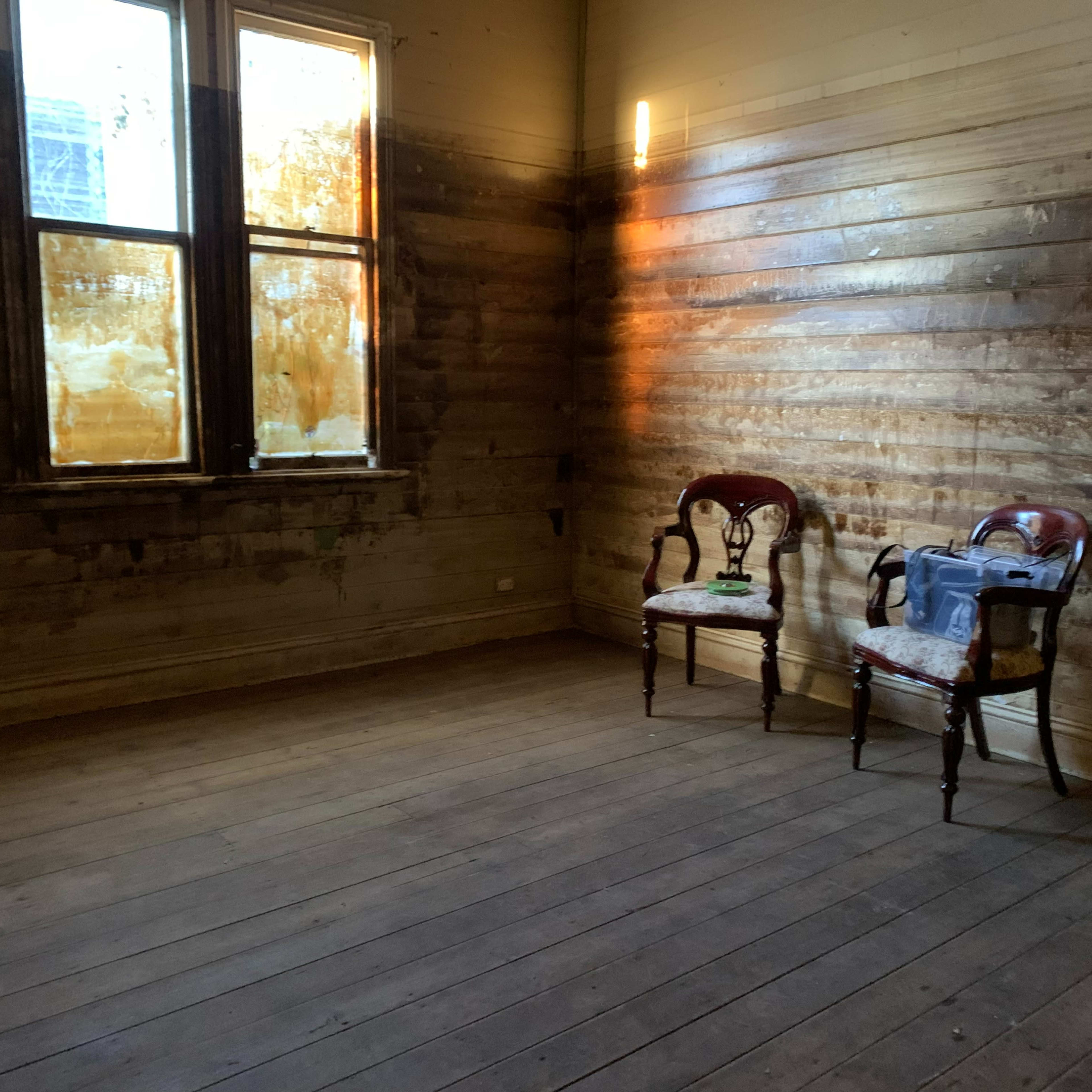 An empty room with two wooden chairs and a bare wooden floor, featuring peeling walls and two windows.