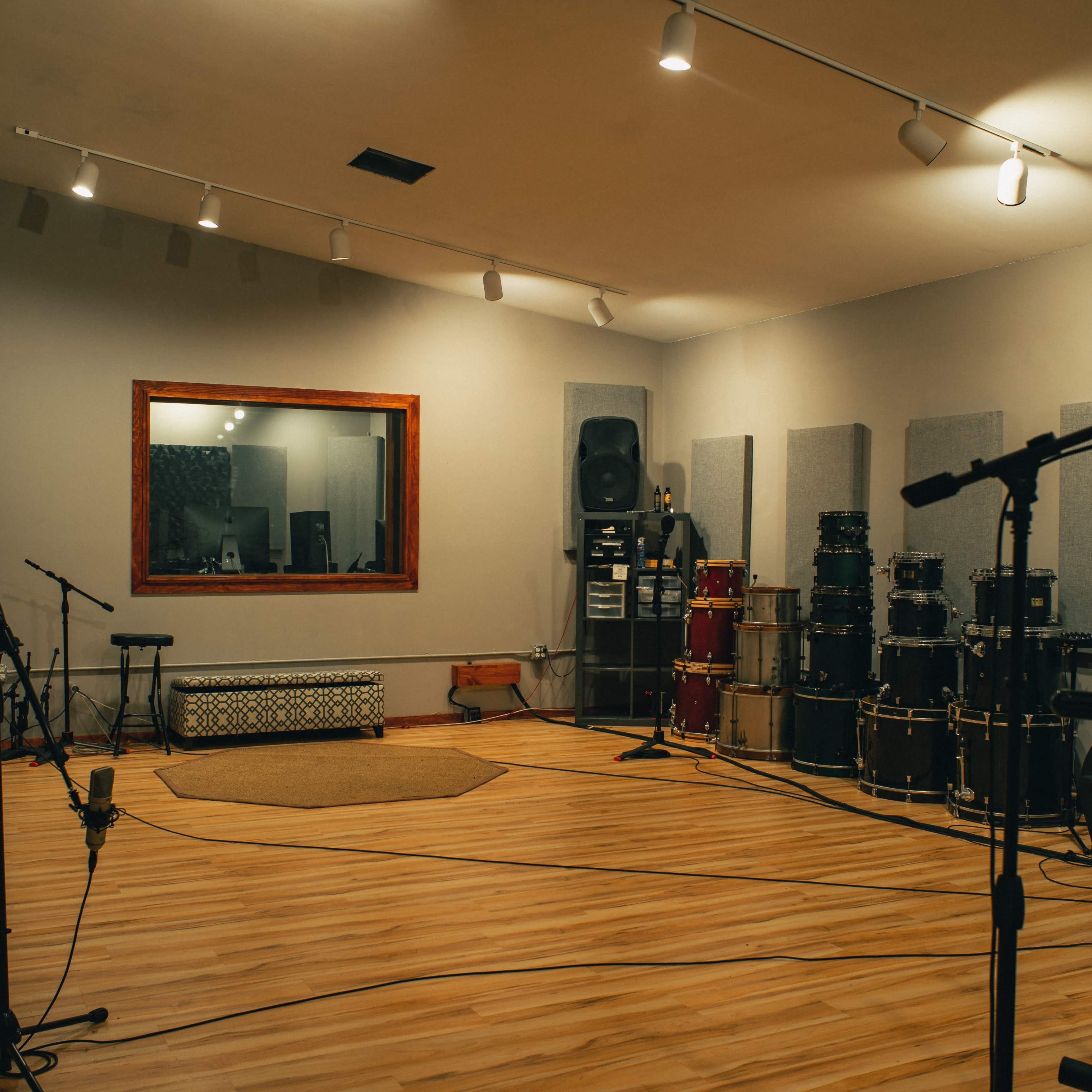 A spacious recording studio featuring various musical instruments, including guitars and a large collection of drums, along with soundproofing panels on the walls.