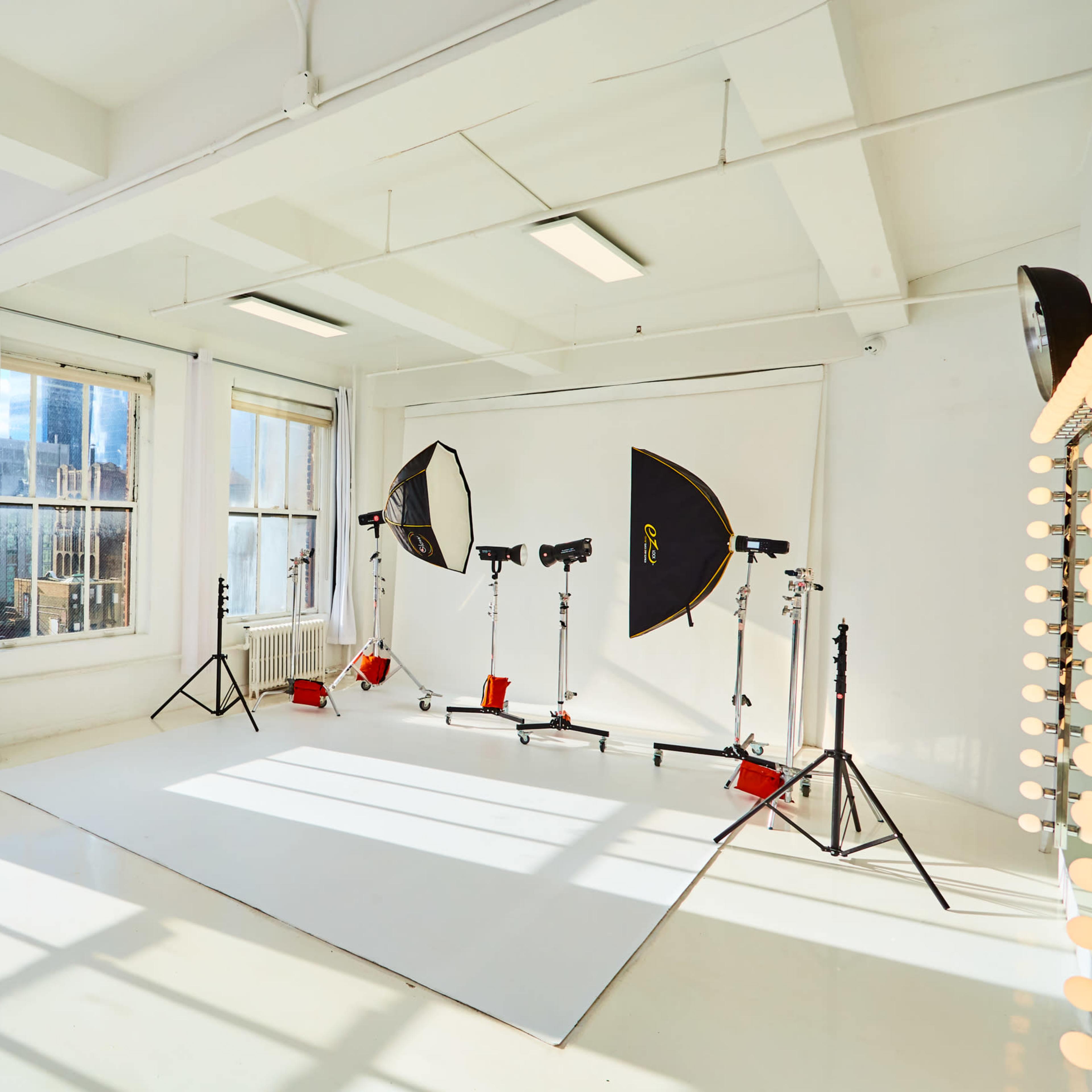 A photography studio with large windows, a seamless white backdrop, and lighting equipment set up on stands.