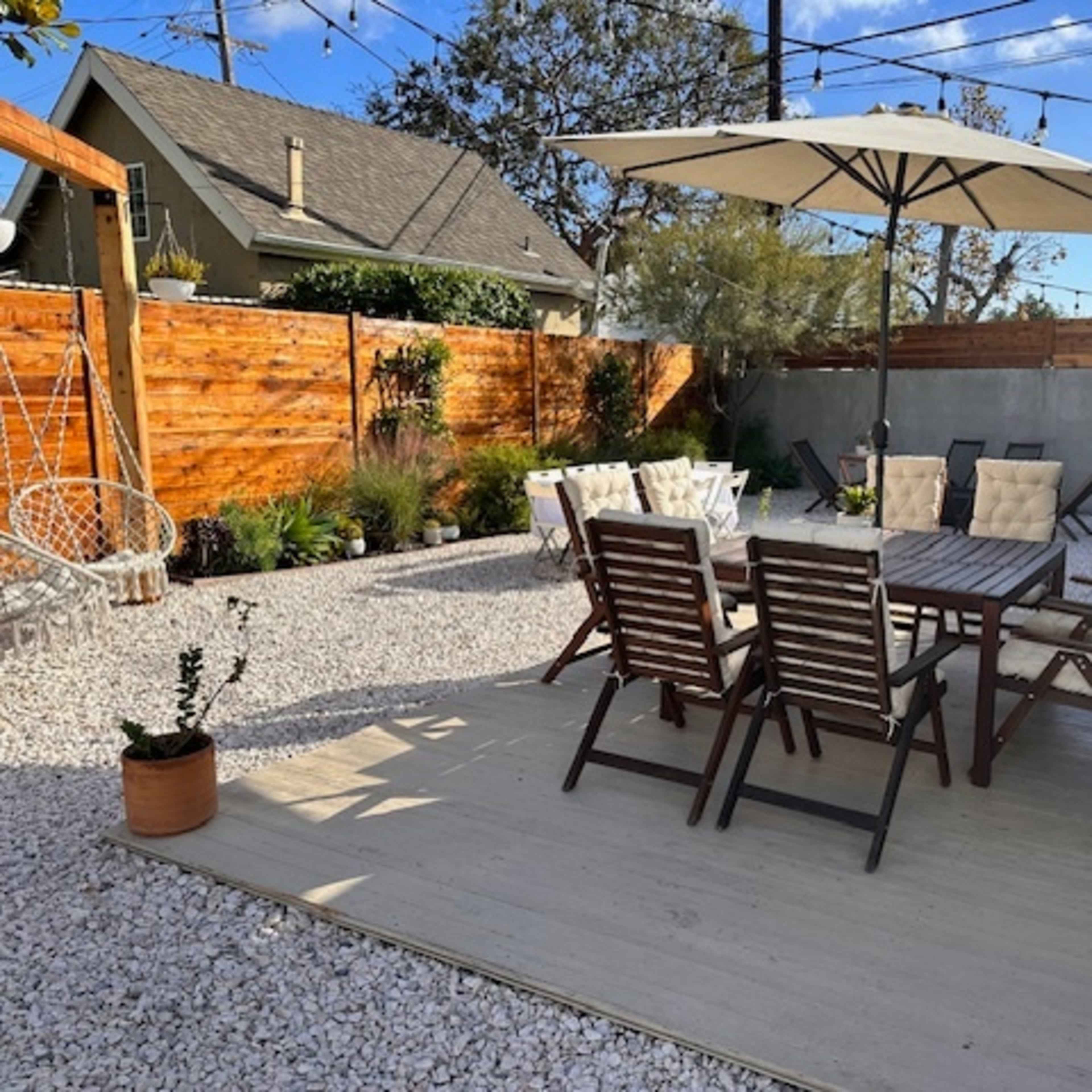 A patio area features a wooden dining table surrounded by folding chairs, an umbrella for shade, and gravel landscaping with plants and a swinging chair.