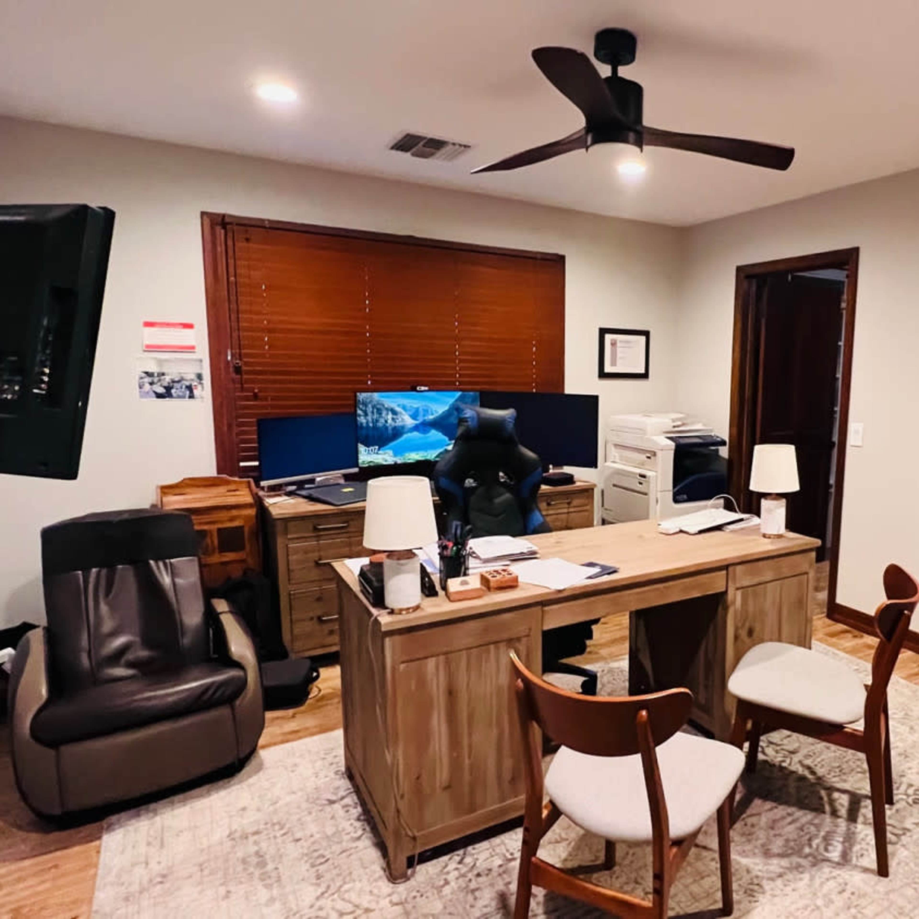 An office with a wooden desk, two chairs, a computer monitor, and various items organized around the space.