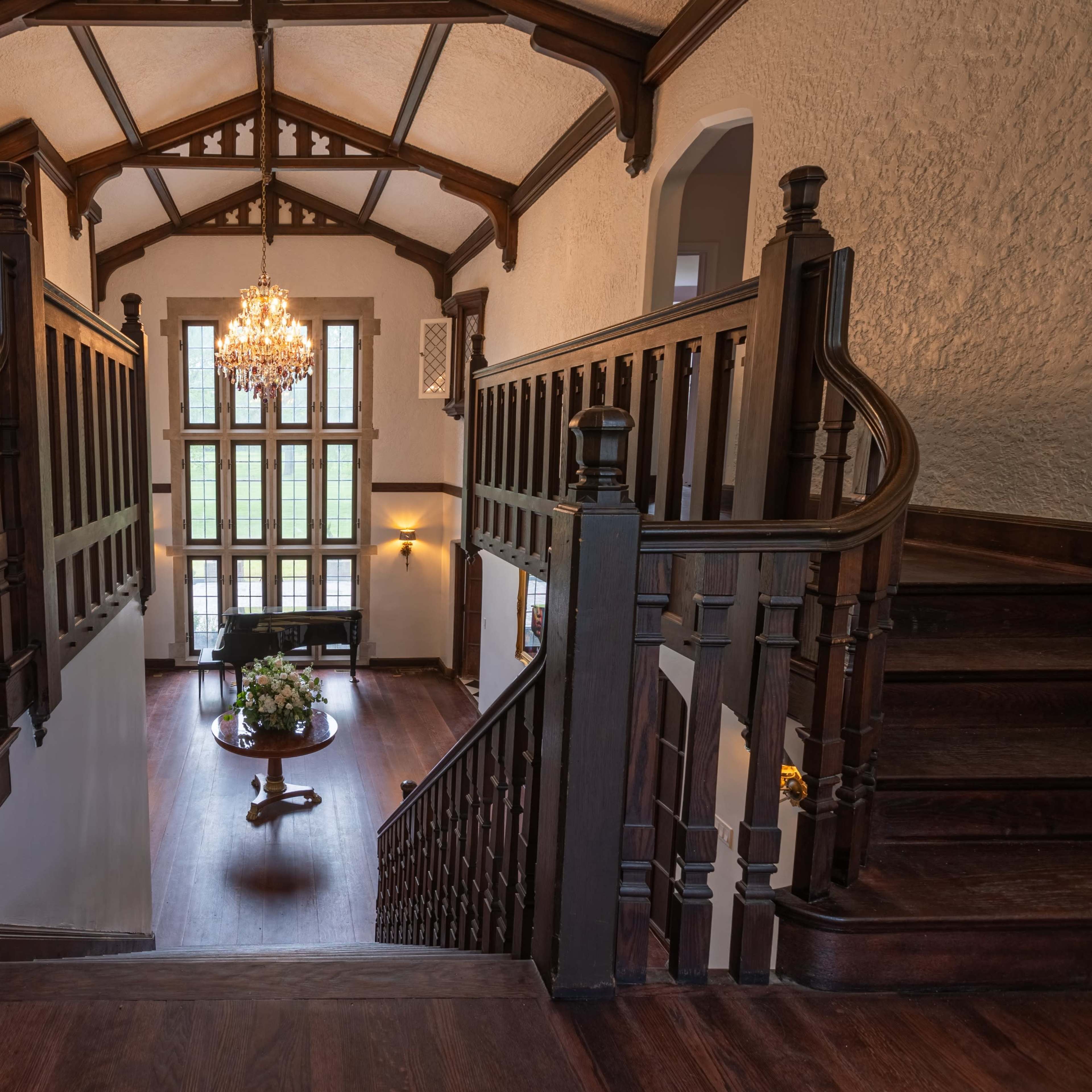 A grand staircase descends from a high ceilinged hallway into a spacious foyer with large windows and elegant decor.