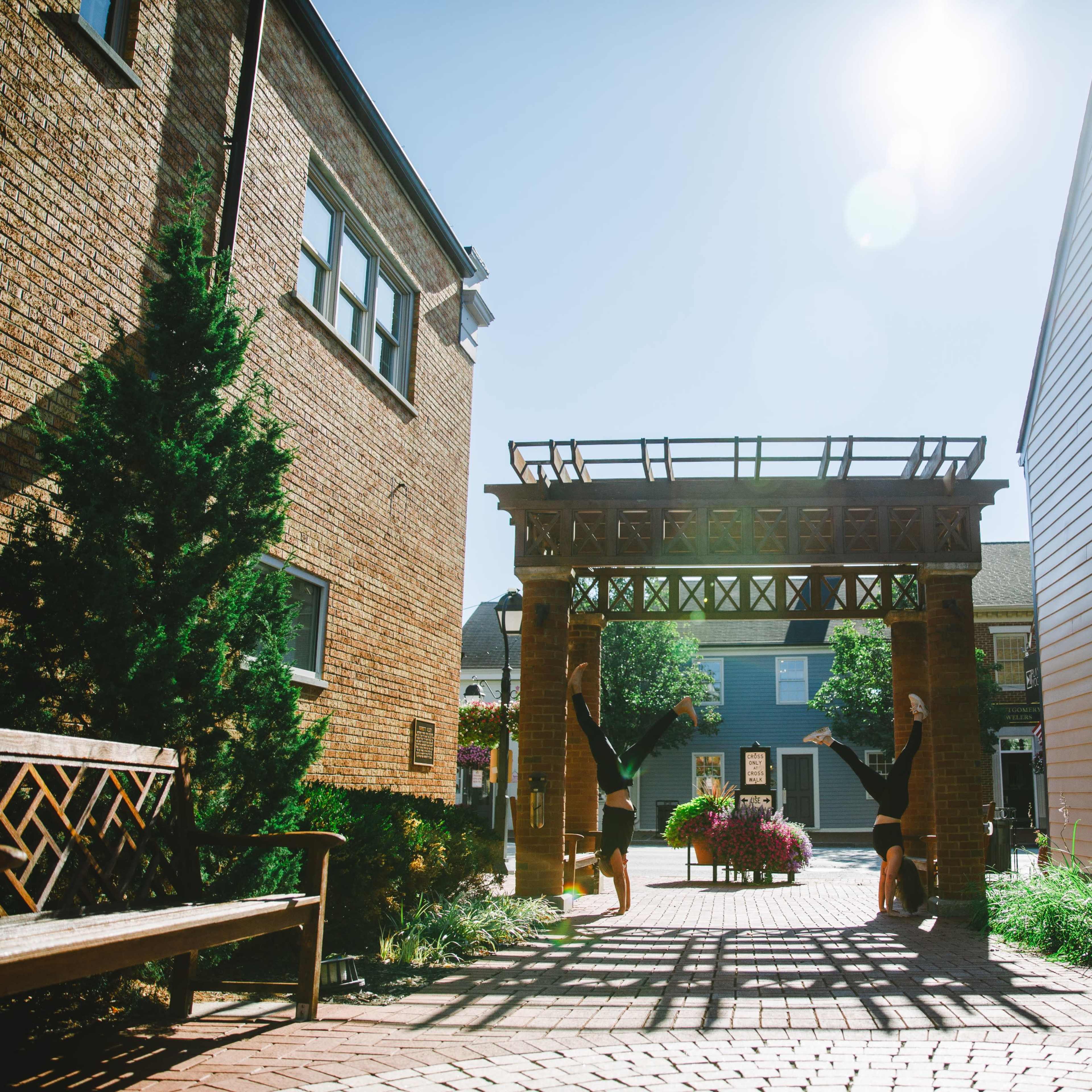 A sunny courtyard with a wooden archway flanked by brick and wooden buildings, and benches on either side.