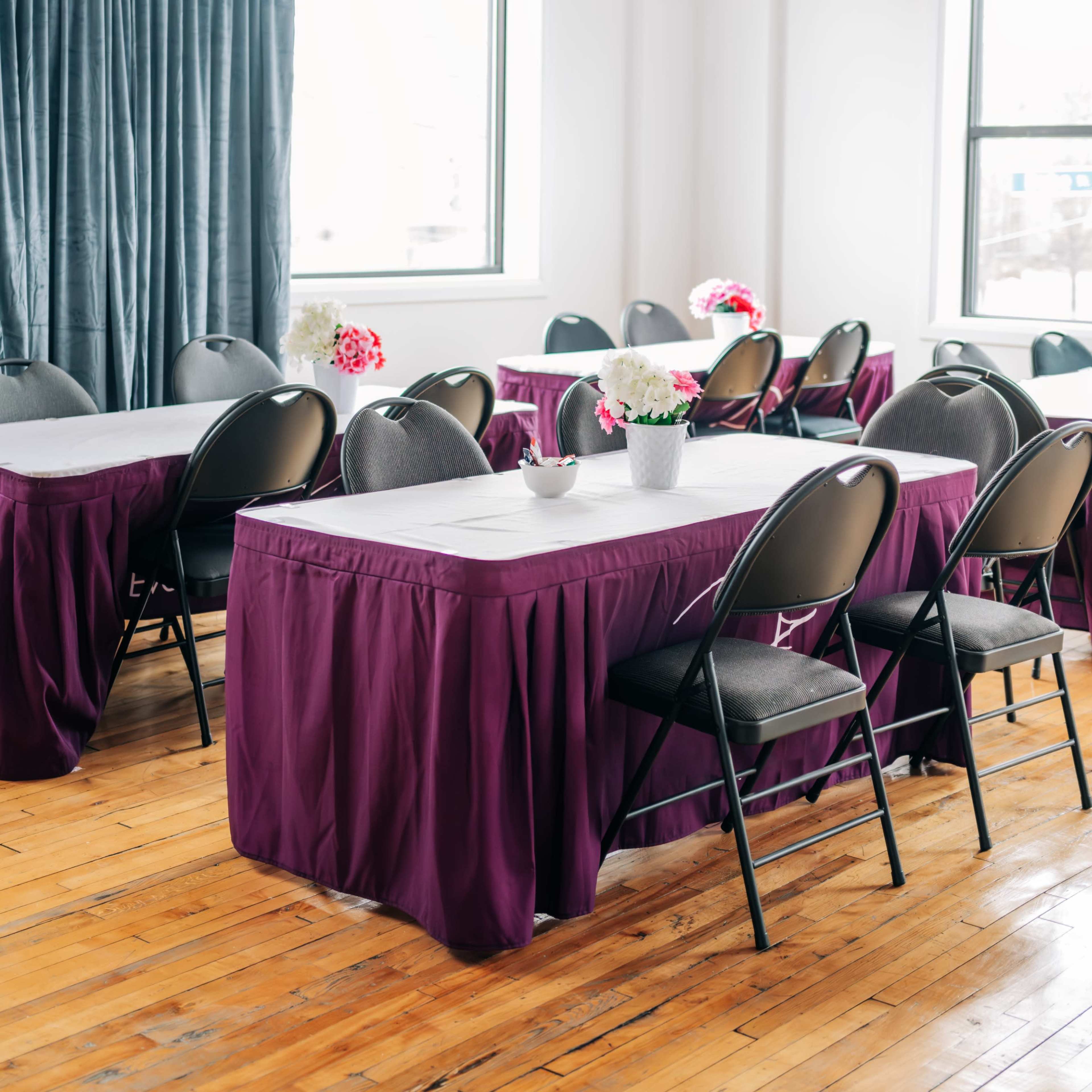 A spacious room features several tables covered with maroon skirts and black chairs, arranged neatly under large windows.
