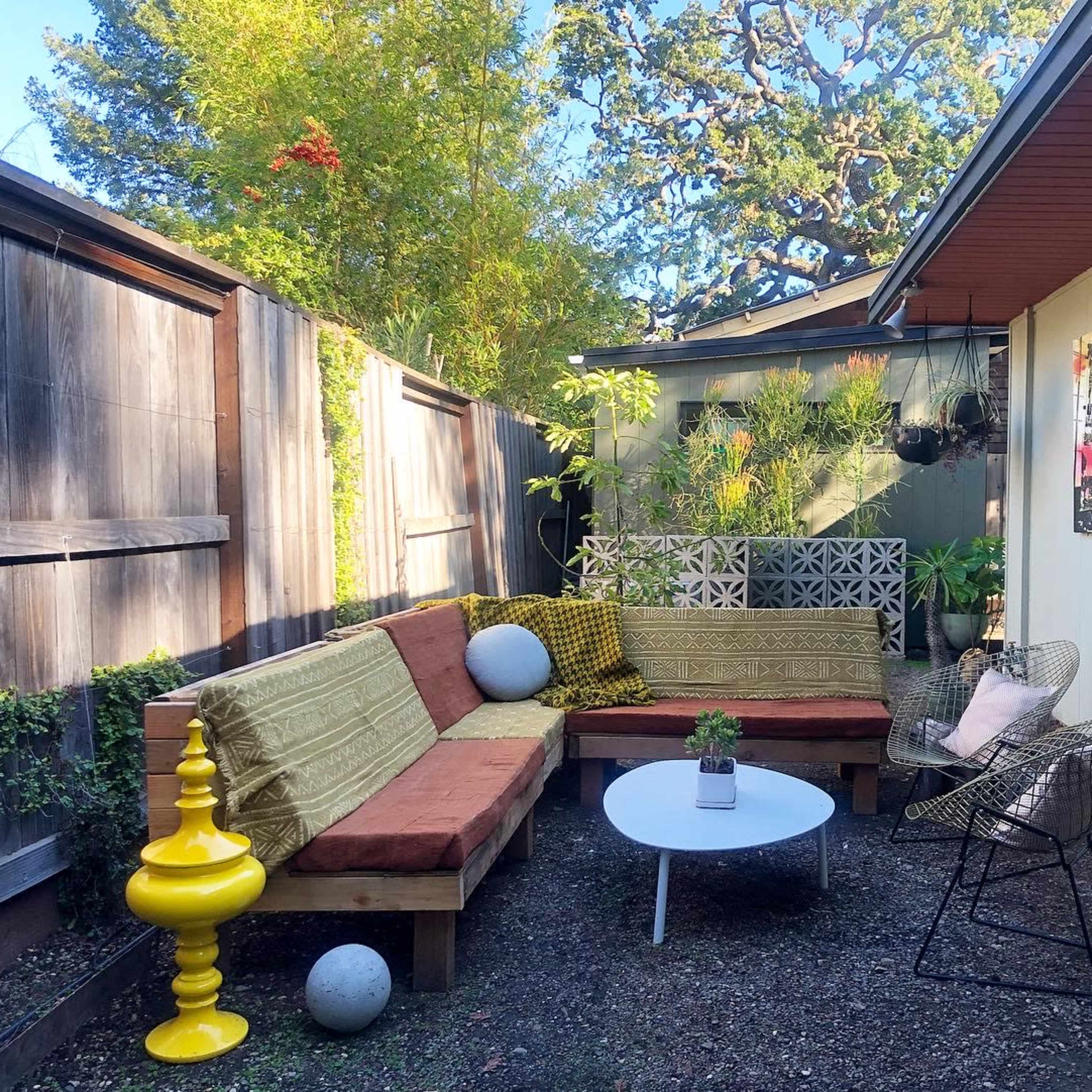 An outdoor seating area with wooden benches covered in cushions, a round white coffee table, and surrounding greenery.