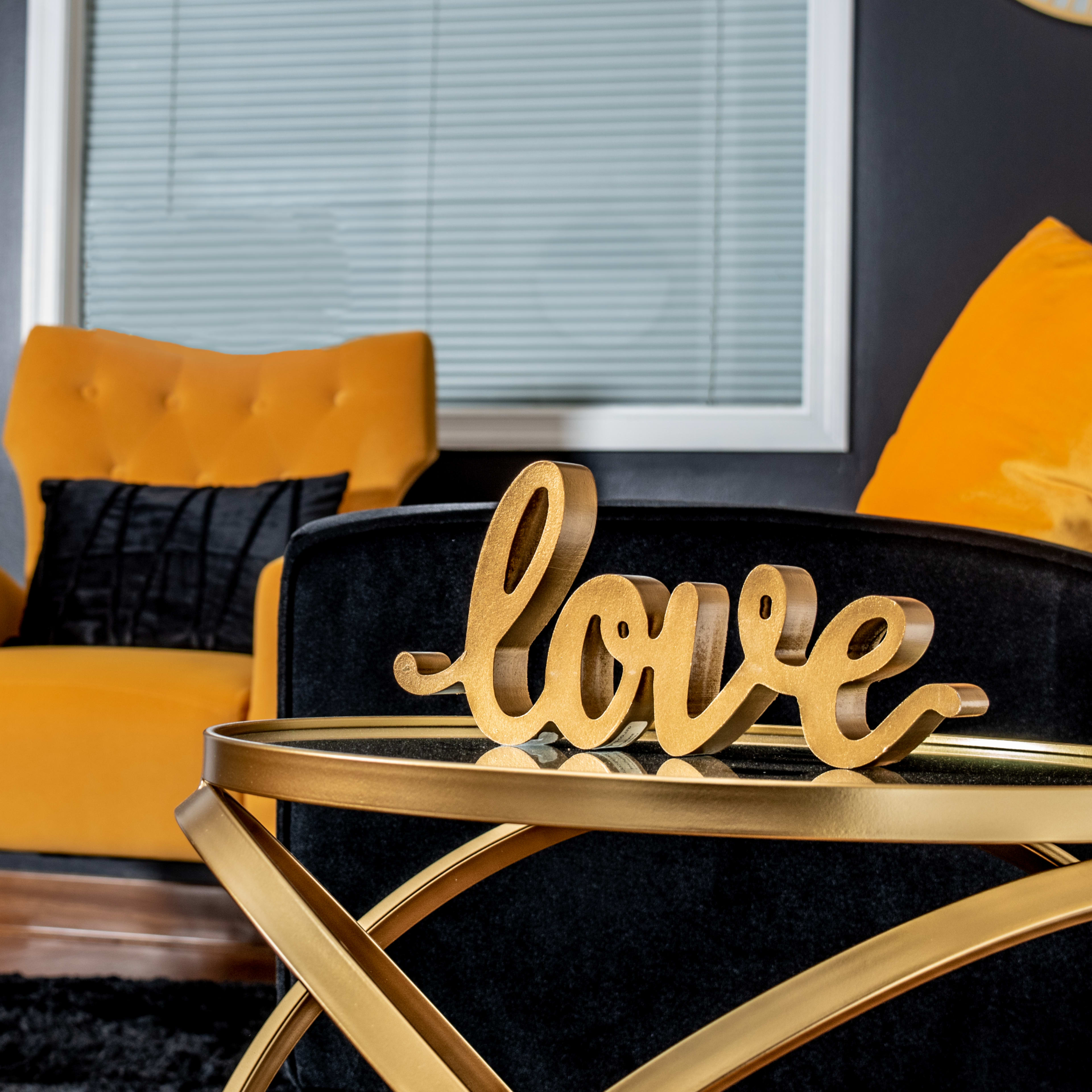 A wooden "love" sign rests on a gold-accented table in a room featuring a yellow armchair and black decor.