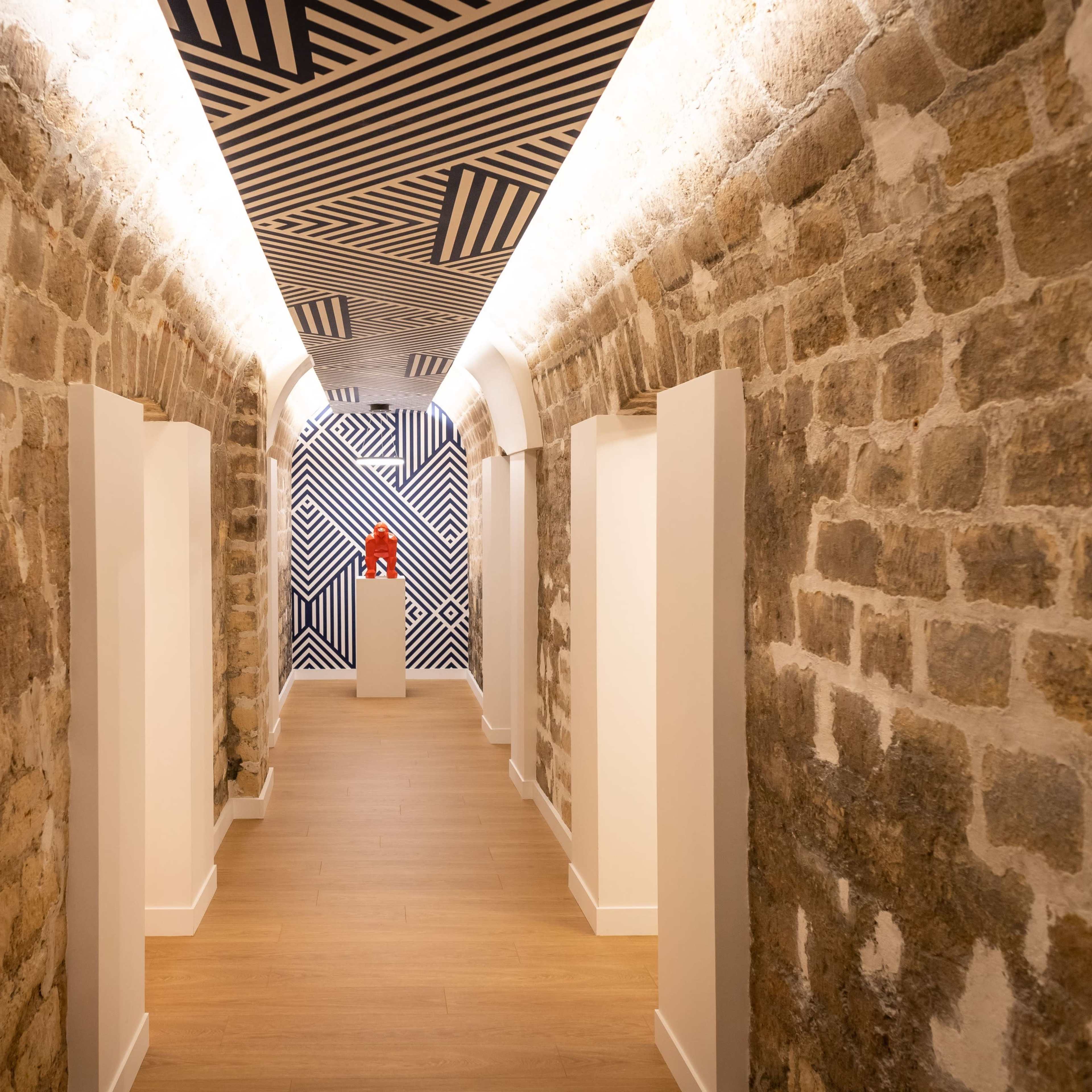 A corridor featuring exposed stone walls, a patterned ceiling, and wooden flooring, with white doors lining the sides.
