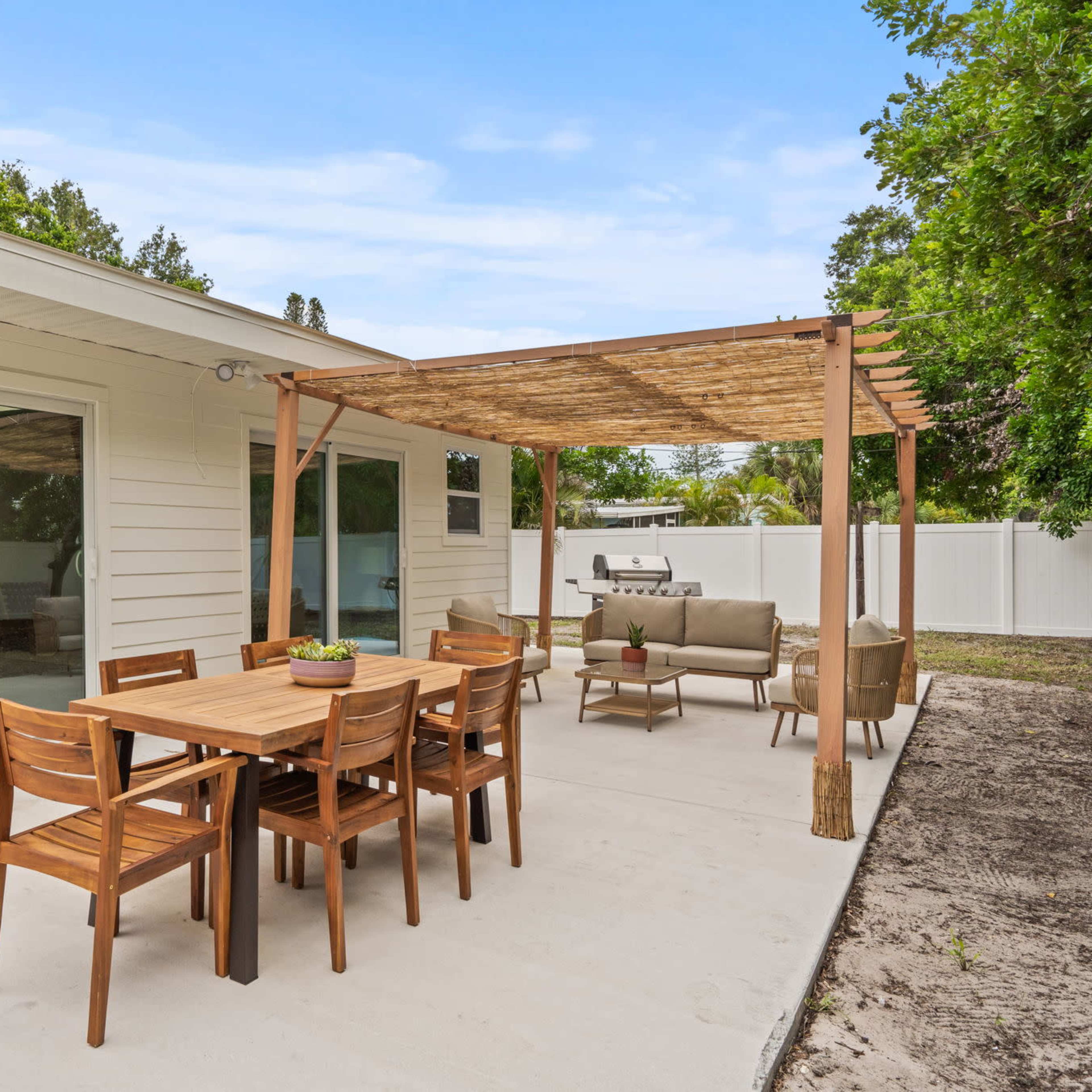 A patio area features a wooden dining set, a seating arrangement under a pergola, and a grill beside a house with large windows.