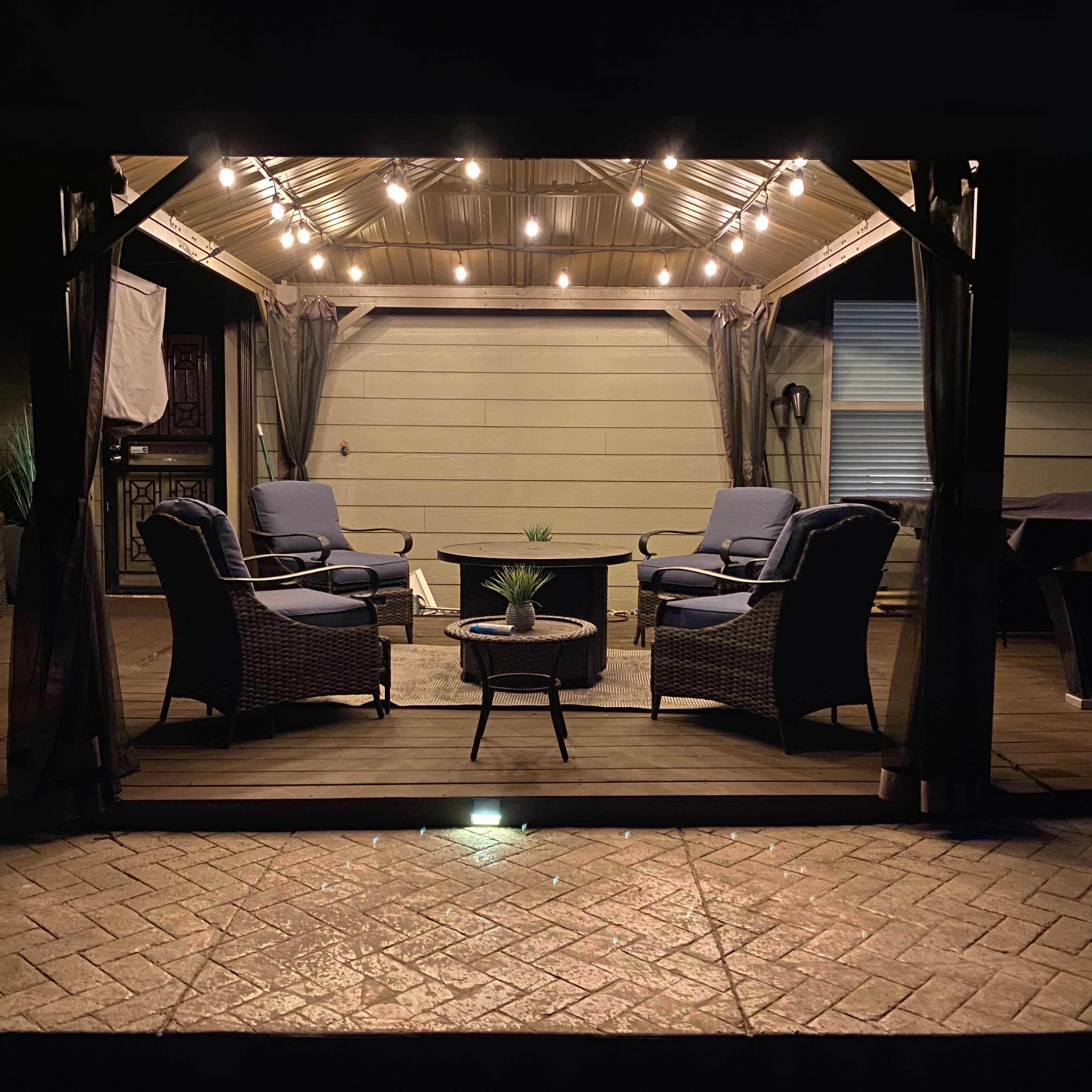 A cozy outdoor seating area is illuminated by string lights under a gazebo, featuring four chairs and a small table.