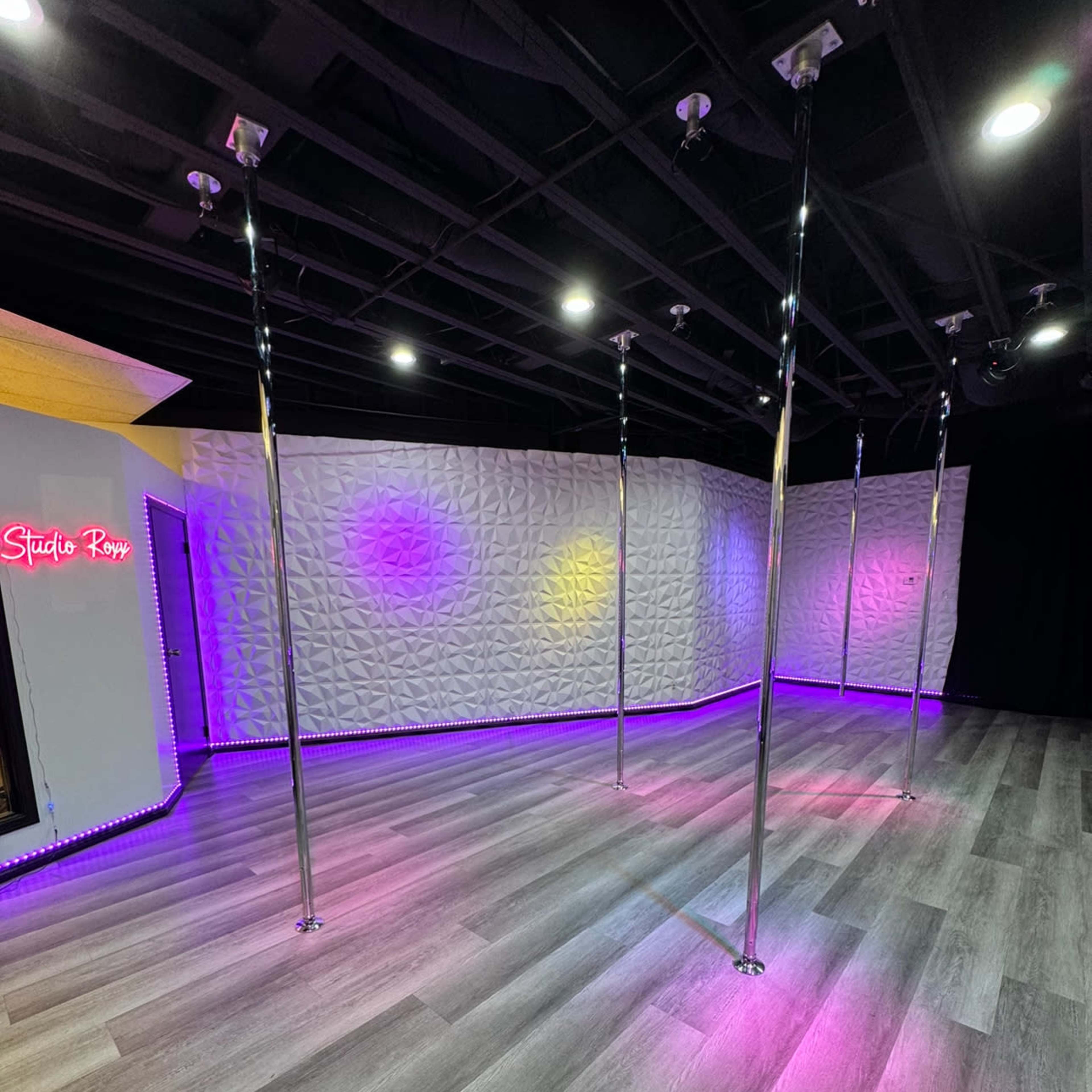 A dance studio features several vertical poles set against a textured wall illuminated by colored lights.