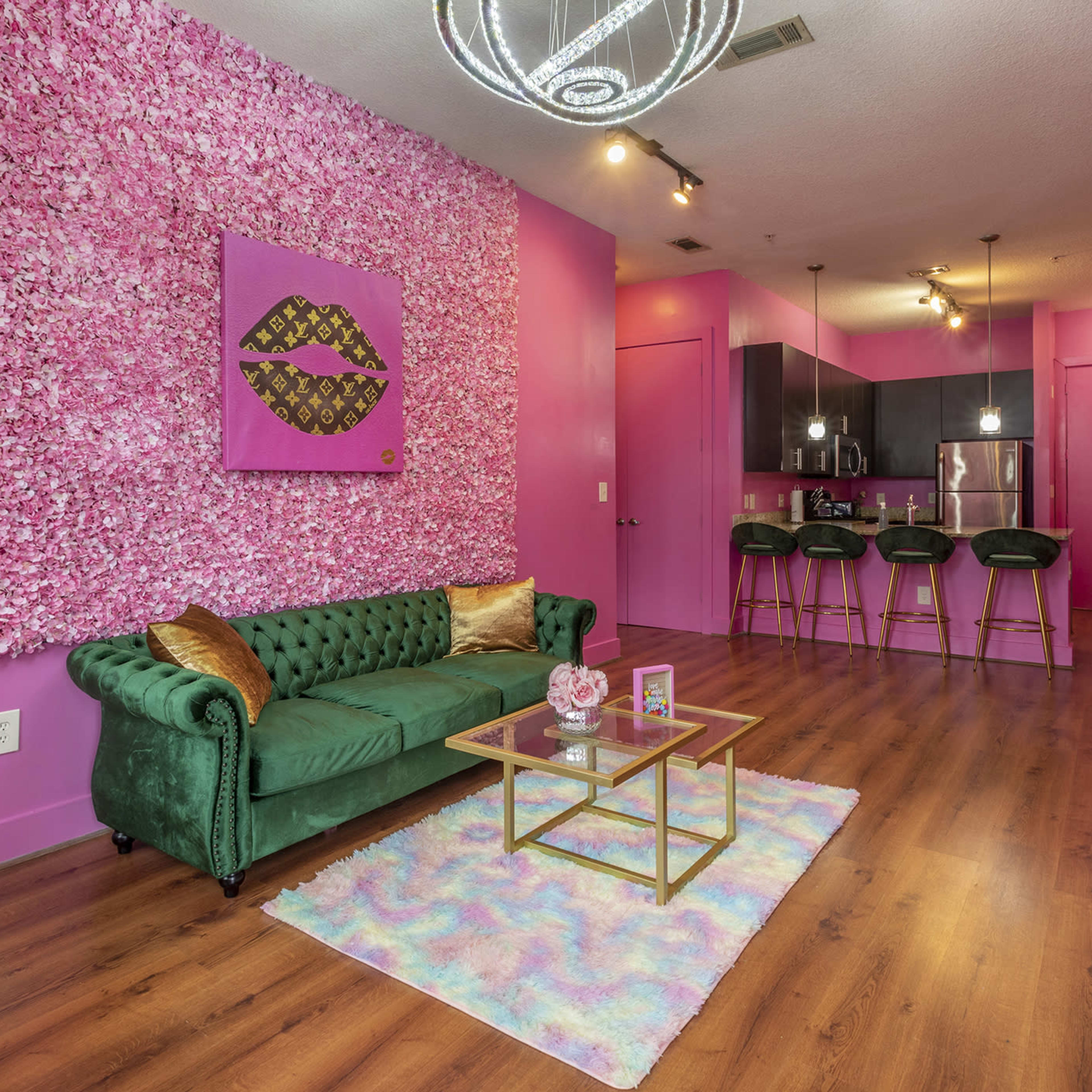 A vibrant living space featuring a green sofa, a wall covered with pink floral decorations, and a small kitchen area in the background.