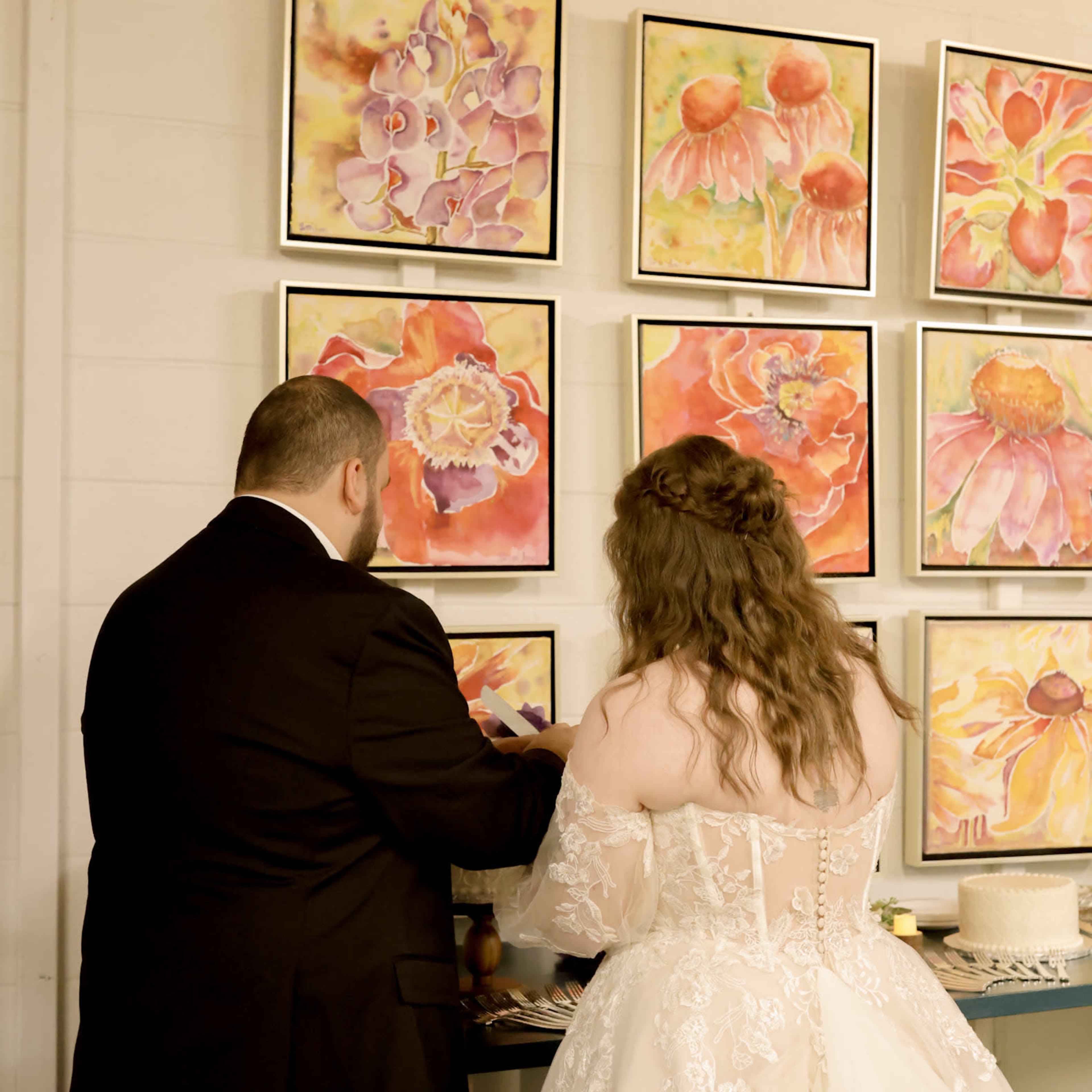 A couple stands together, with the man in a black suit and the woman in a lace wedding dress, facing a wall adorned with colorful floral paintings.