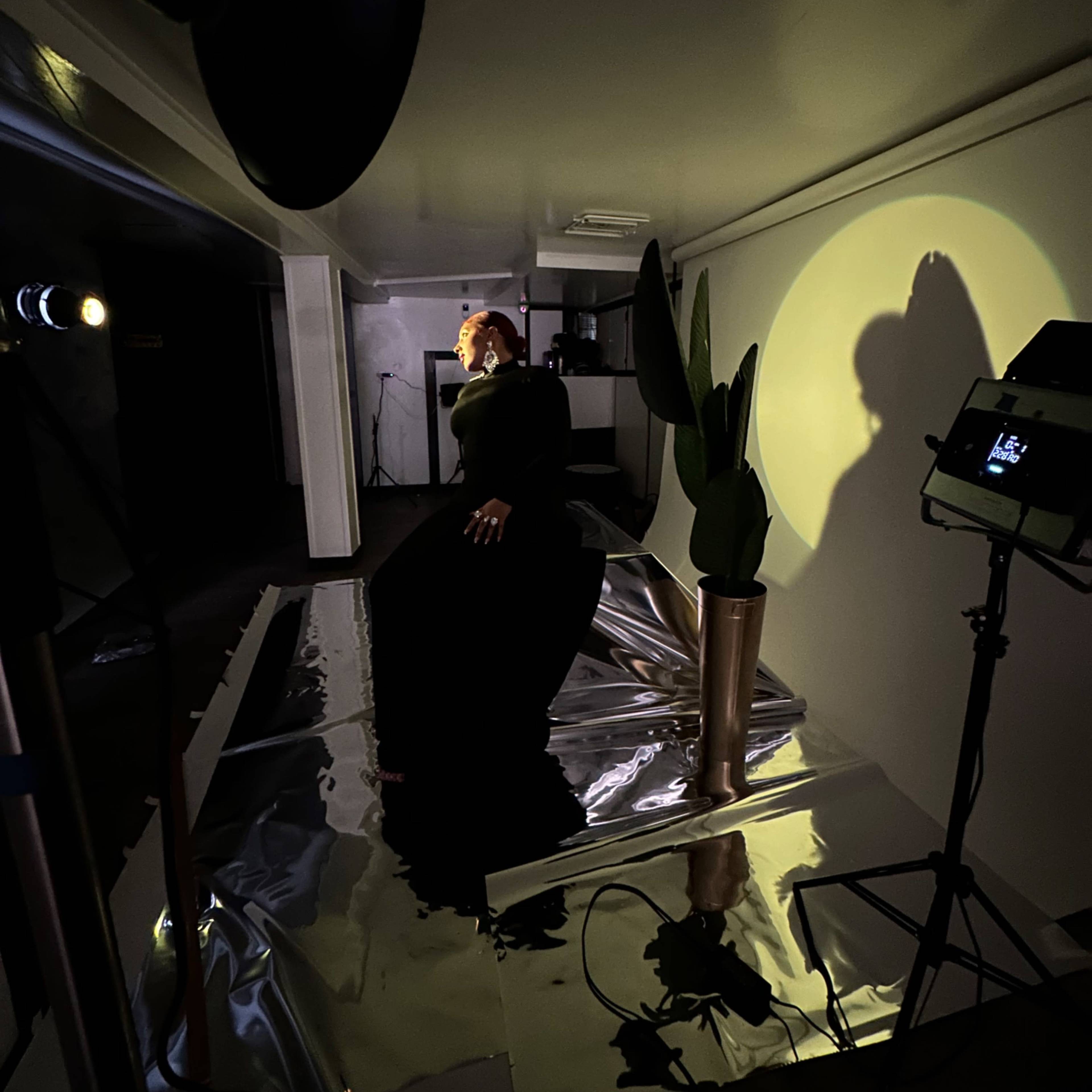 A person in a black outfit sits on a reflective surface next to a potted plant under dim, controlled lighting.
