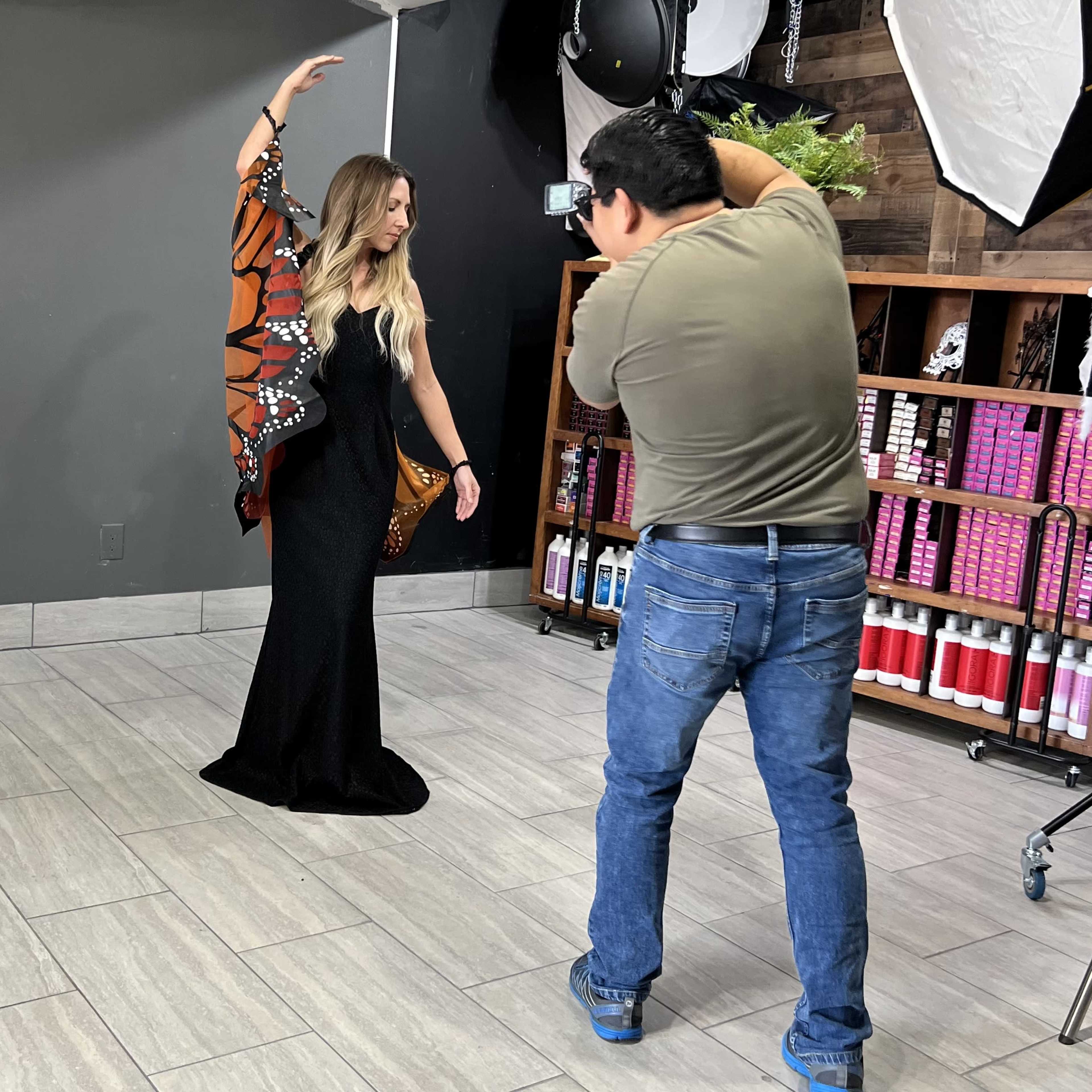 A photographer is capturing an image of a model wearing a black dress and a patterned shawl in a studio setting.