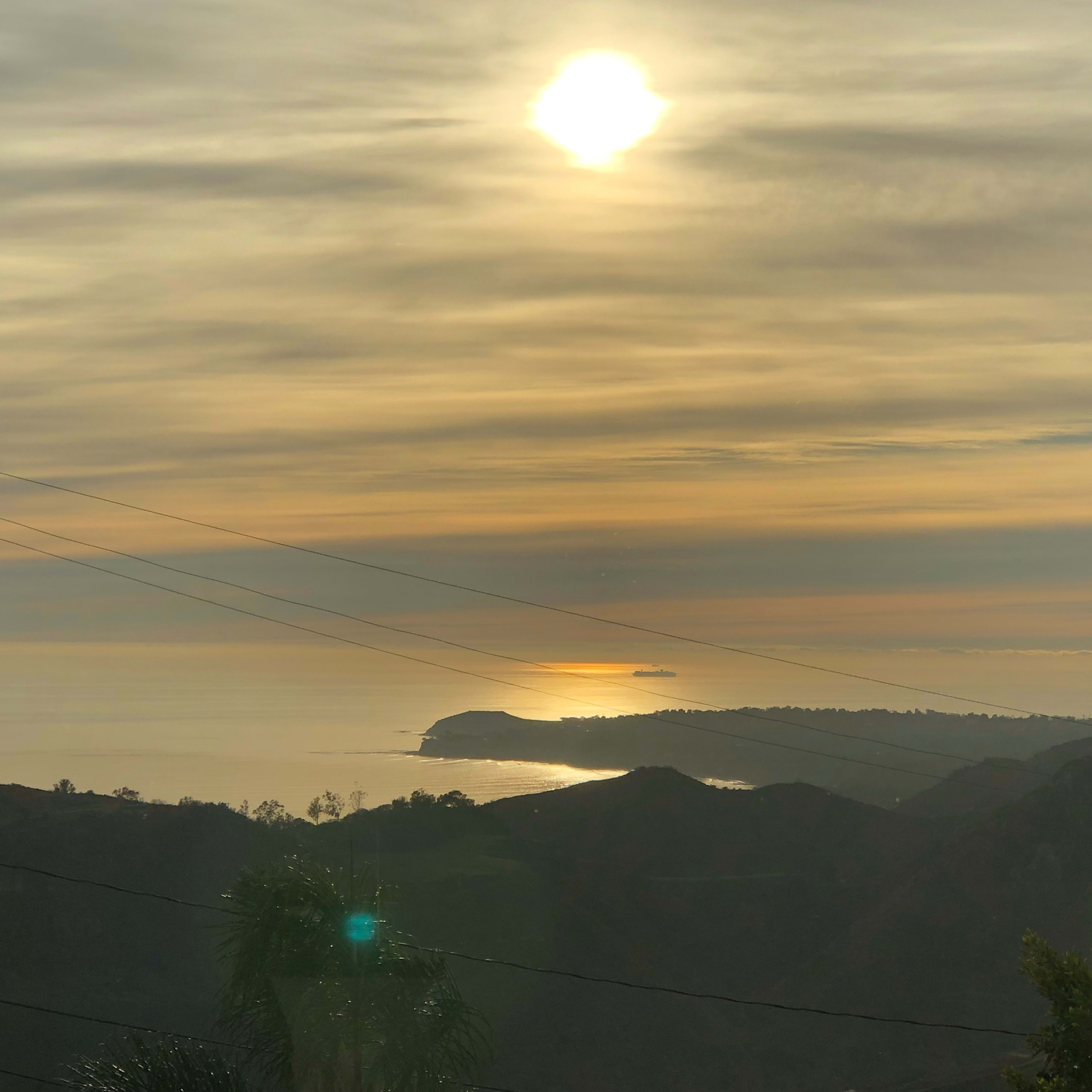 A sunset over a calm ocean, with a faint outline of a distant shoreline and silhouetted hills in the foreground.
