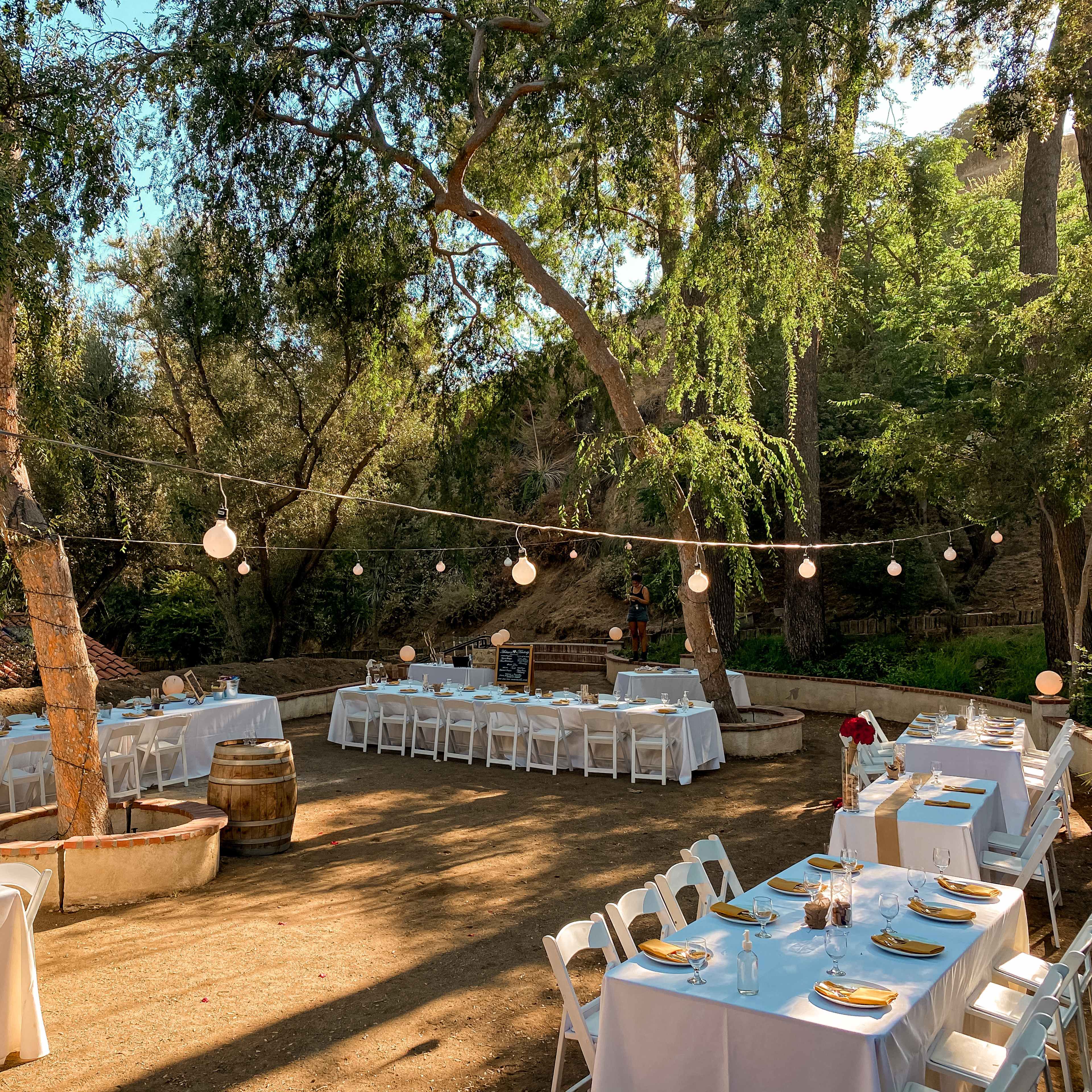 A dining area with white tables and chairs is set up outdoors under trees, adorned with string lights.