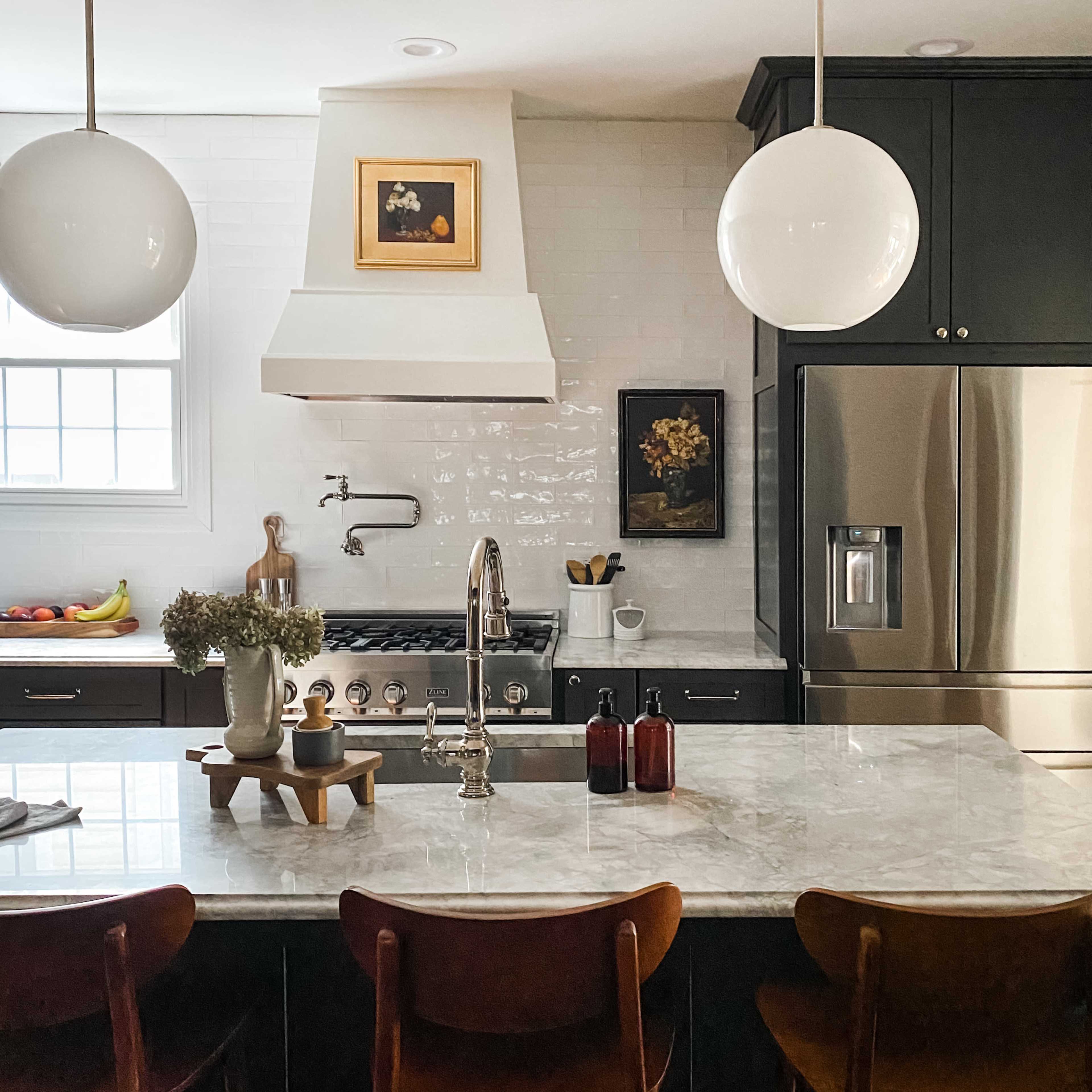 A modern kitchen features a marble island with three wooden stools, stainless steel appliances, and a white backsplash with decorative framed art.