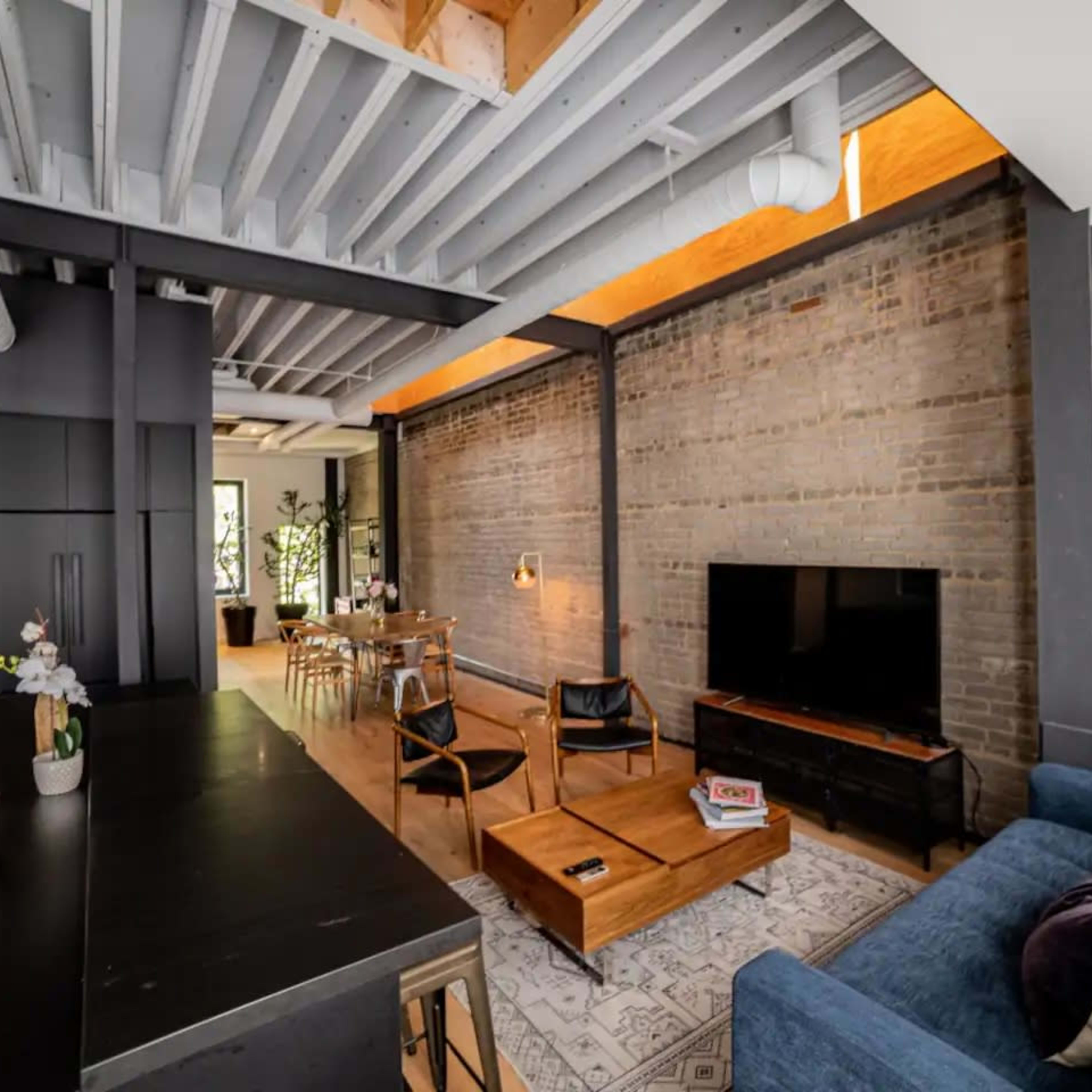An open living space with a modern kitchen, dining area, and a comfortable seating arrangement, featuring exposed brick walls and wooden beams.