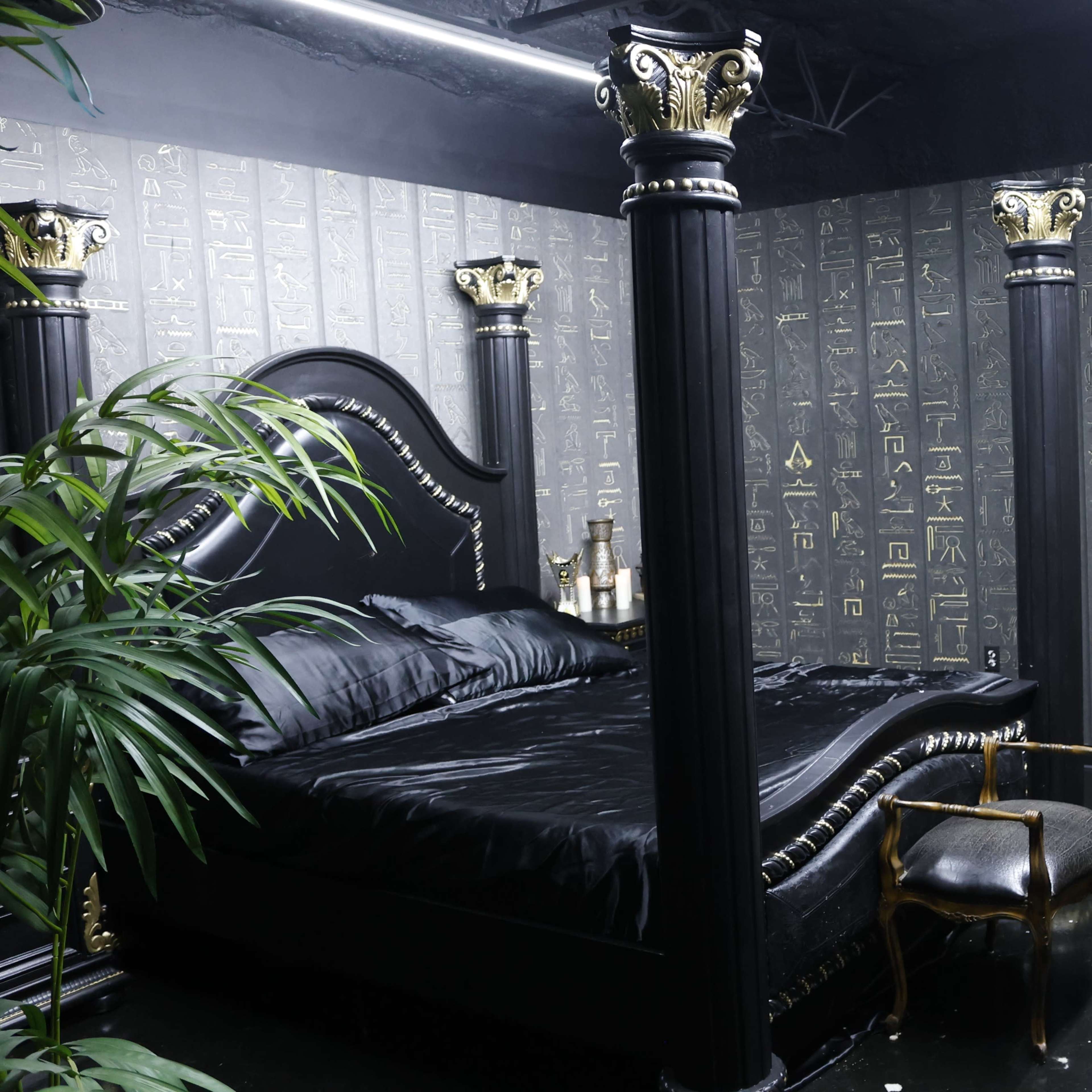 A dark room featuring a black bed with ornate columns and walls decorated with hieroglyphics.