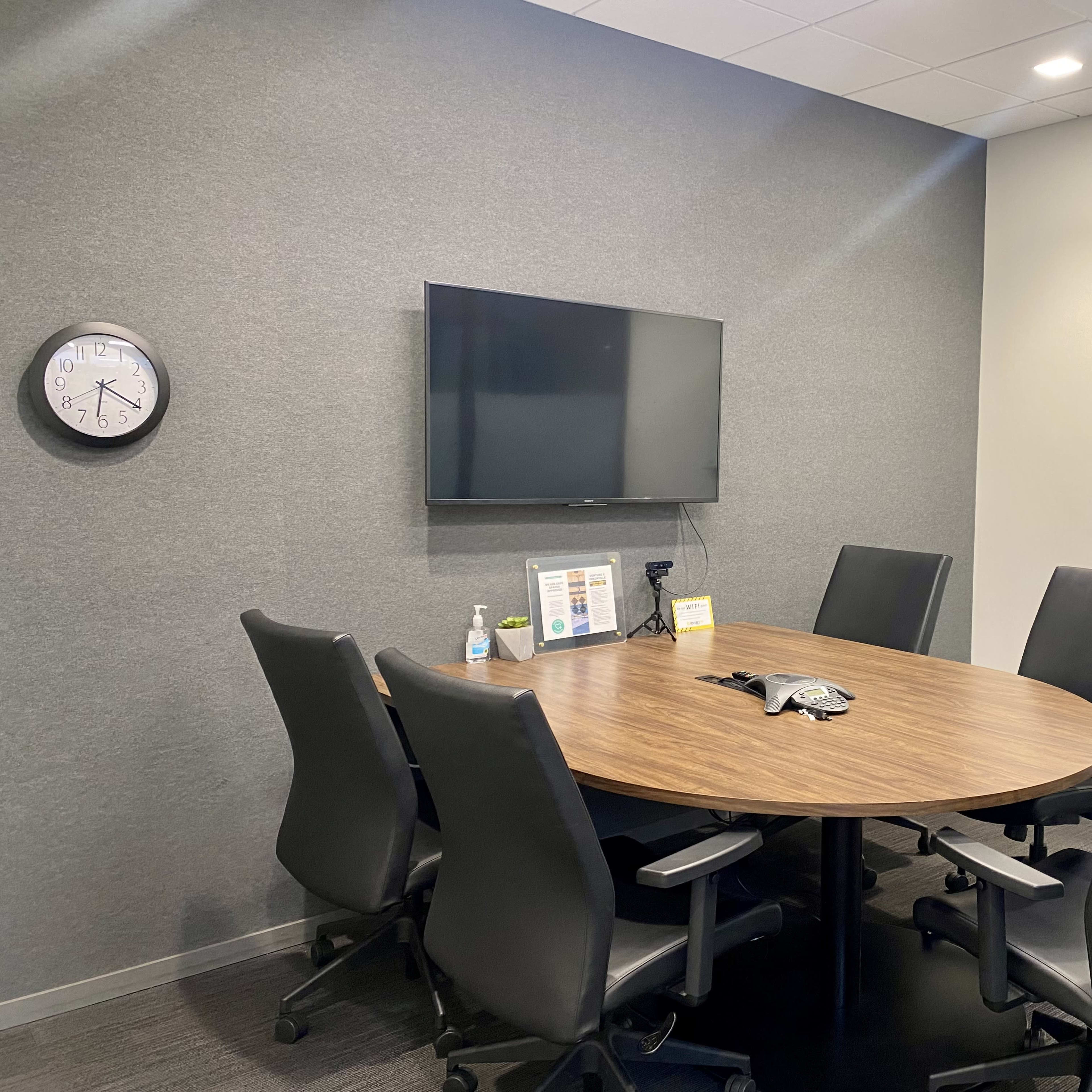 A modern conference room with a round wooden table surrounded by six black ergonomic chairs, a wall-mounted TV, a clock, and a whiteboard.