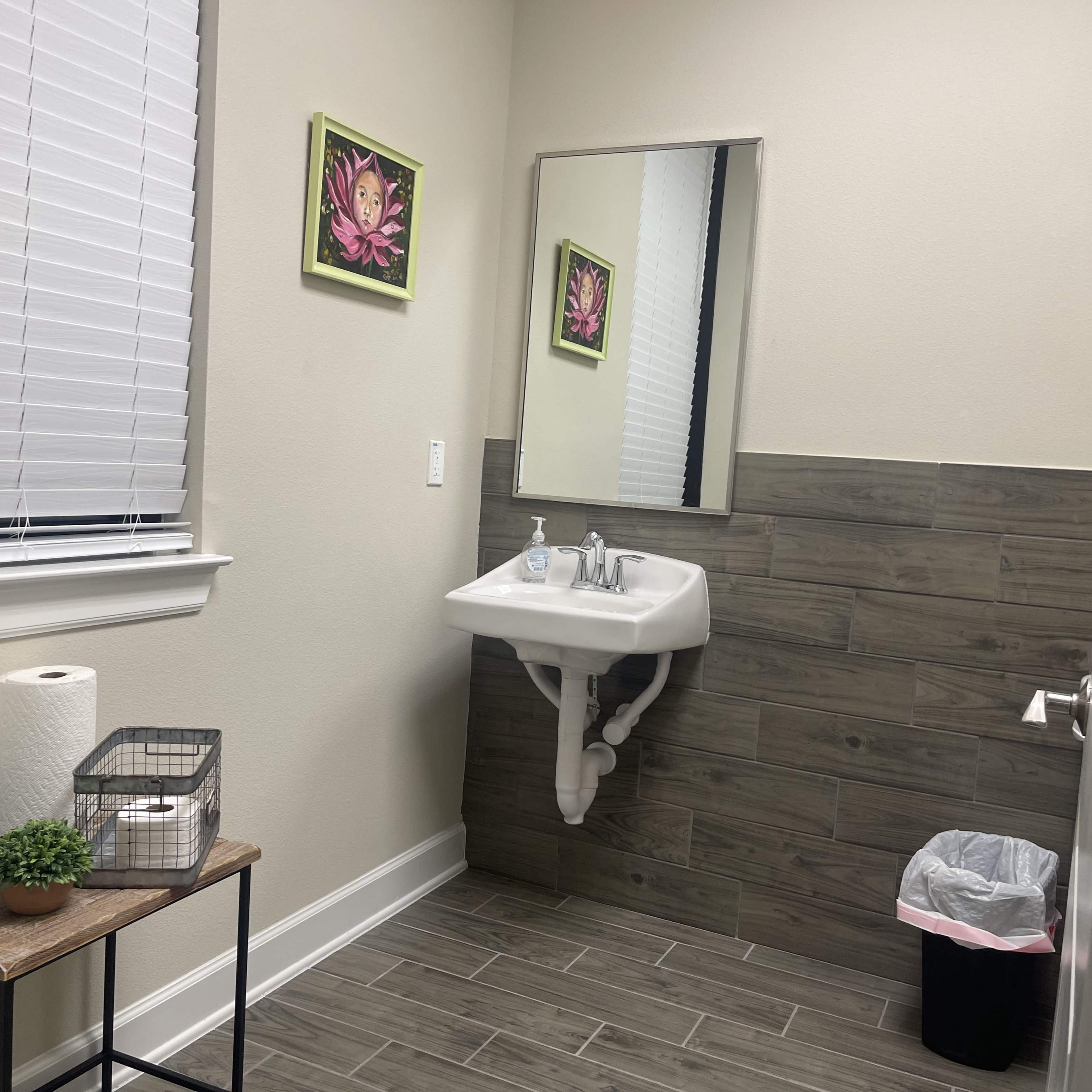 A small bathroom with a wall-mounted sink, a mirror, a trash can, and decorative artwork on the walls.