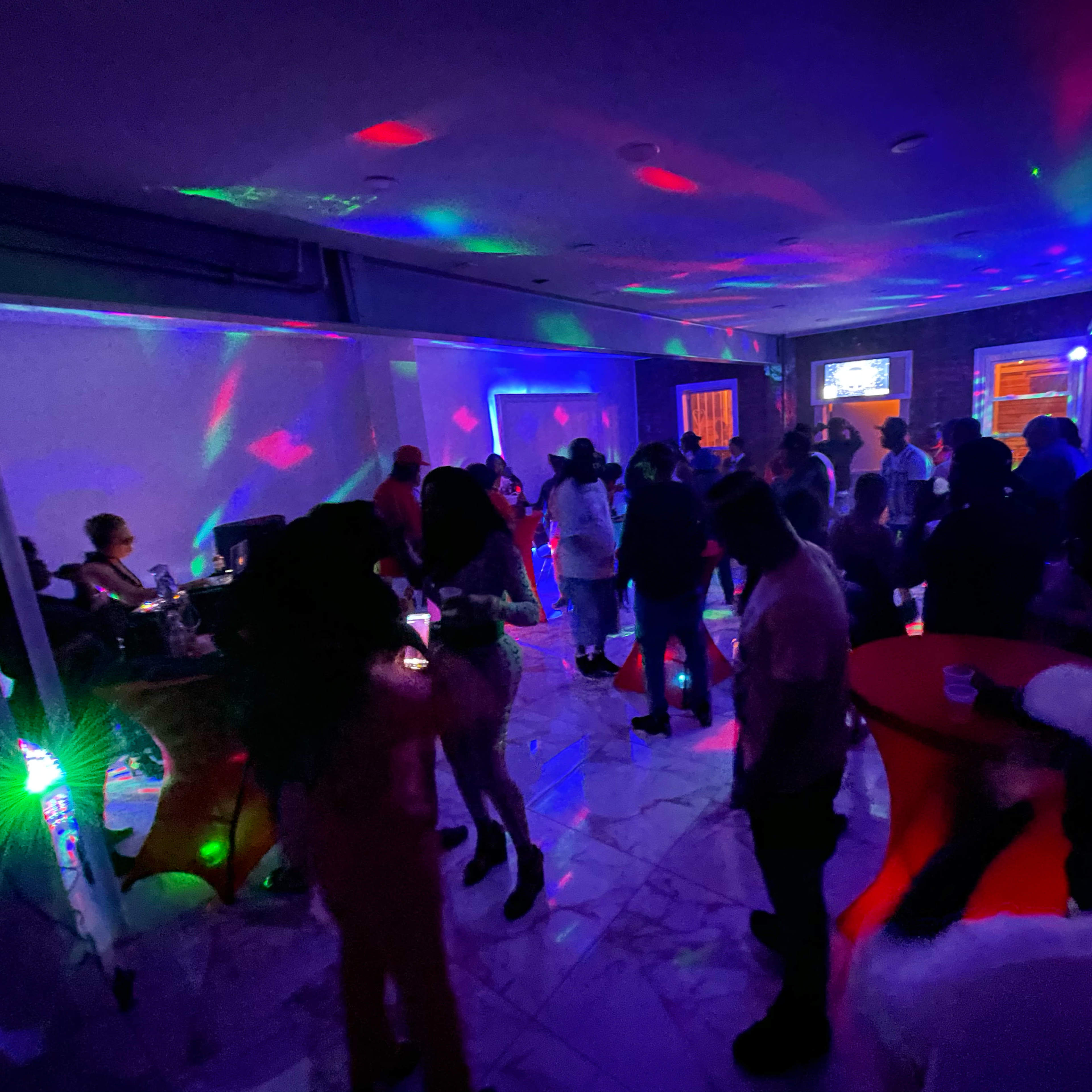 A crowded event space features colorful lights and music as people socialize and dance.