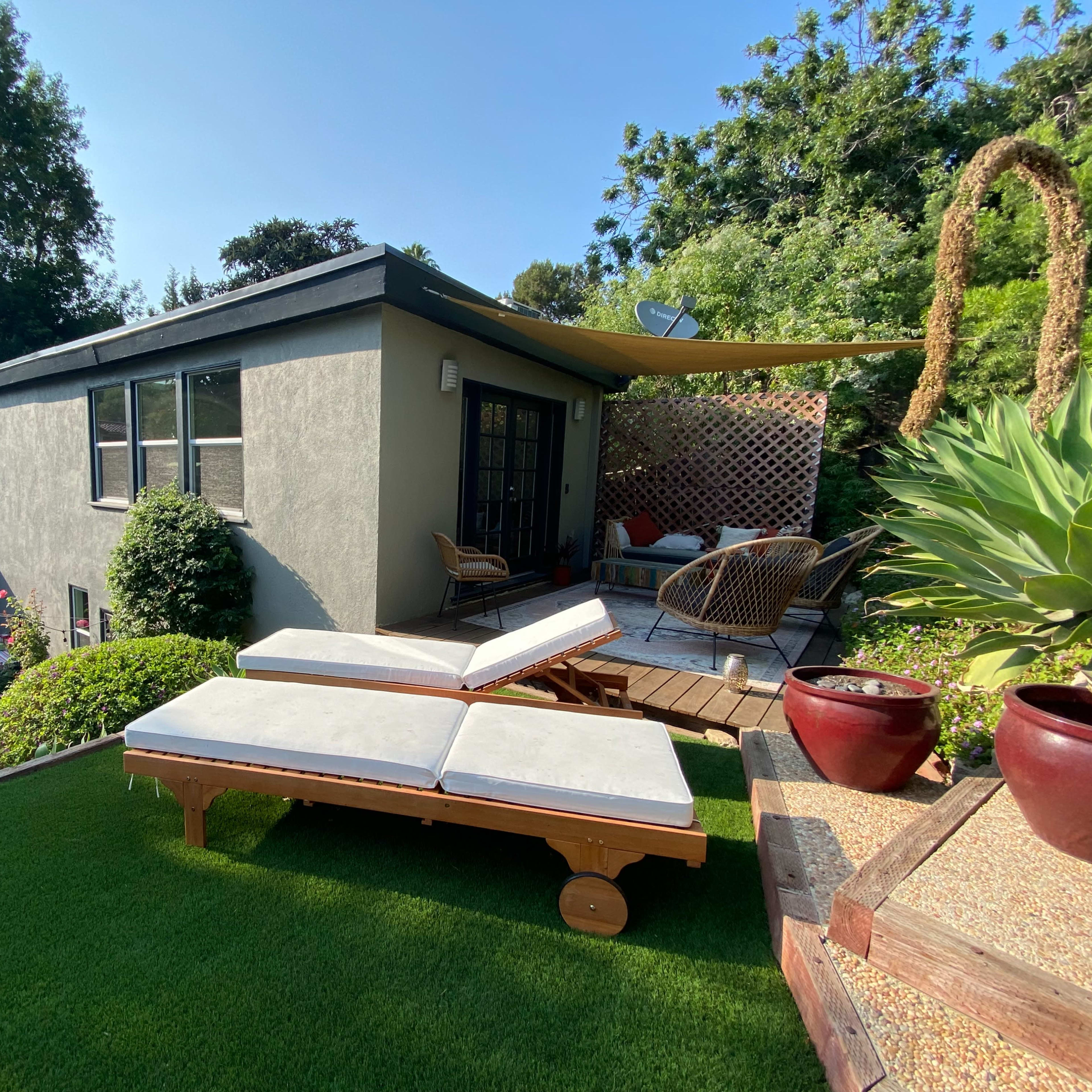 A backyard with two lounge chairs on artificial grass, a small patio area with seating, and a house surrounded by greenery.