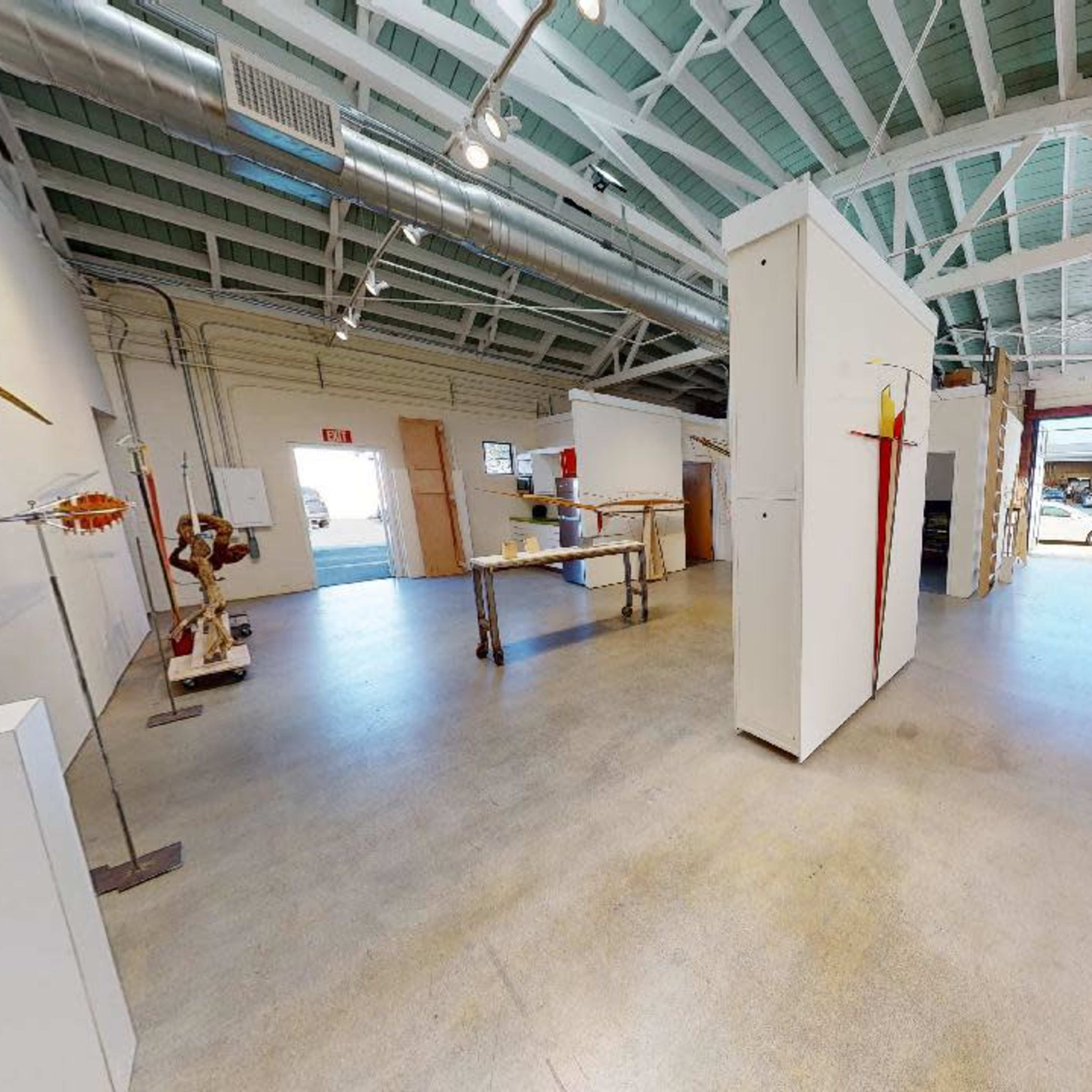 A spacious, well-lit art gallery with white walls and a concrete floor, featuring various sculptures and art installations.