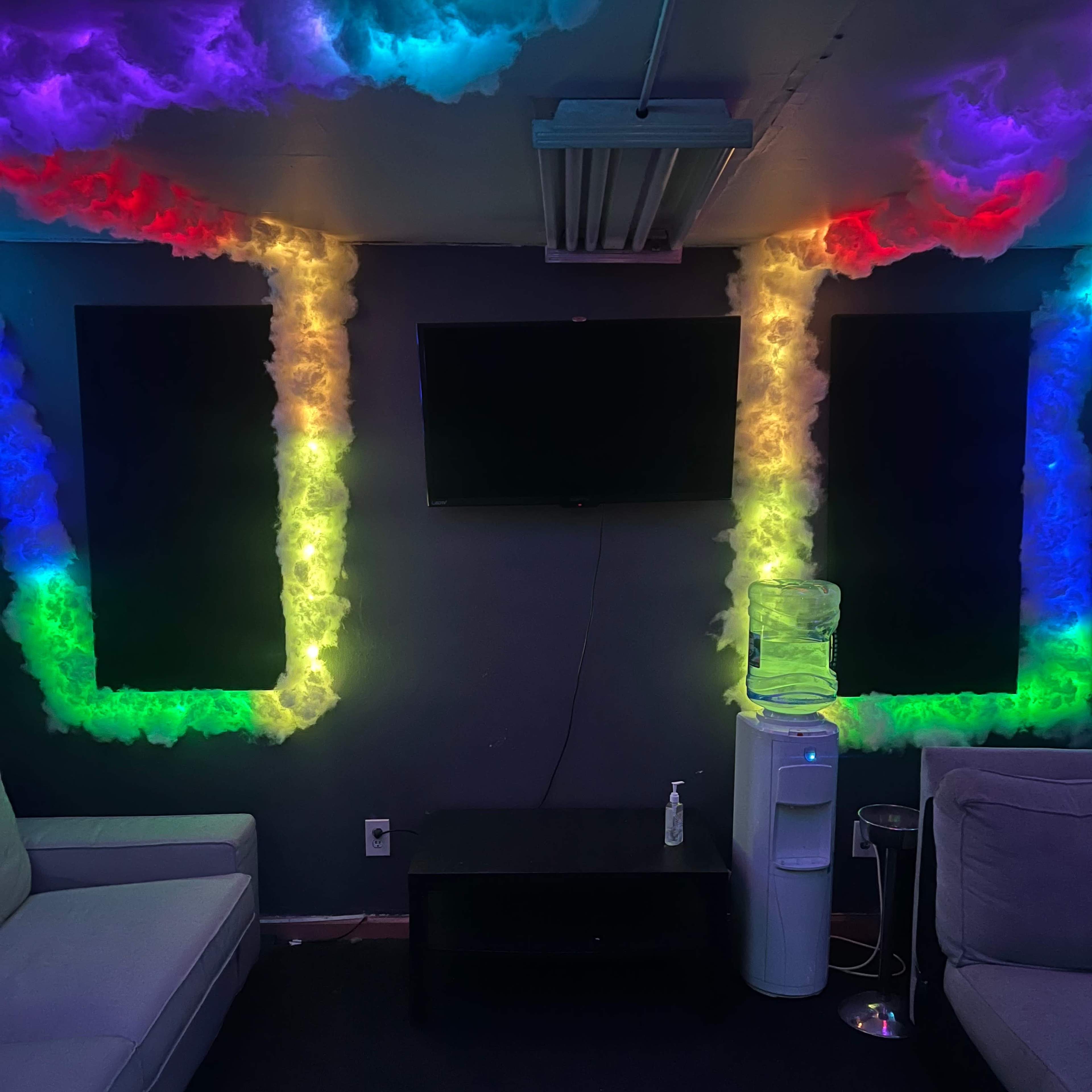 A lounge area with a dark wall adorned with colorful cloud-like light decorations, a television mounted in the center, and a water cooler beside a small black table and light-colored sofas.