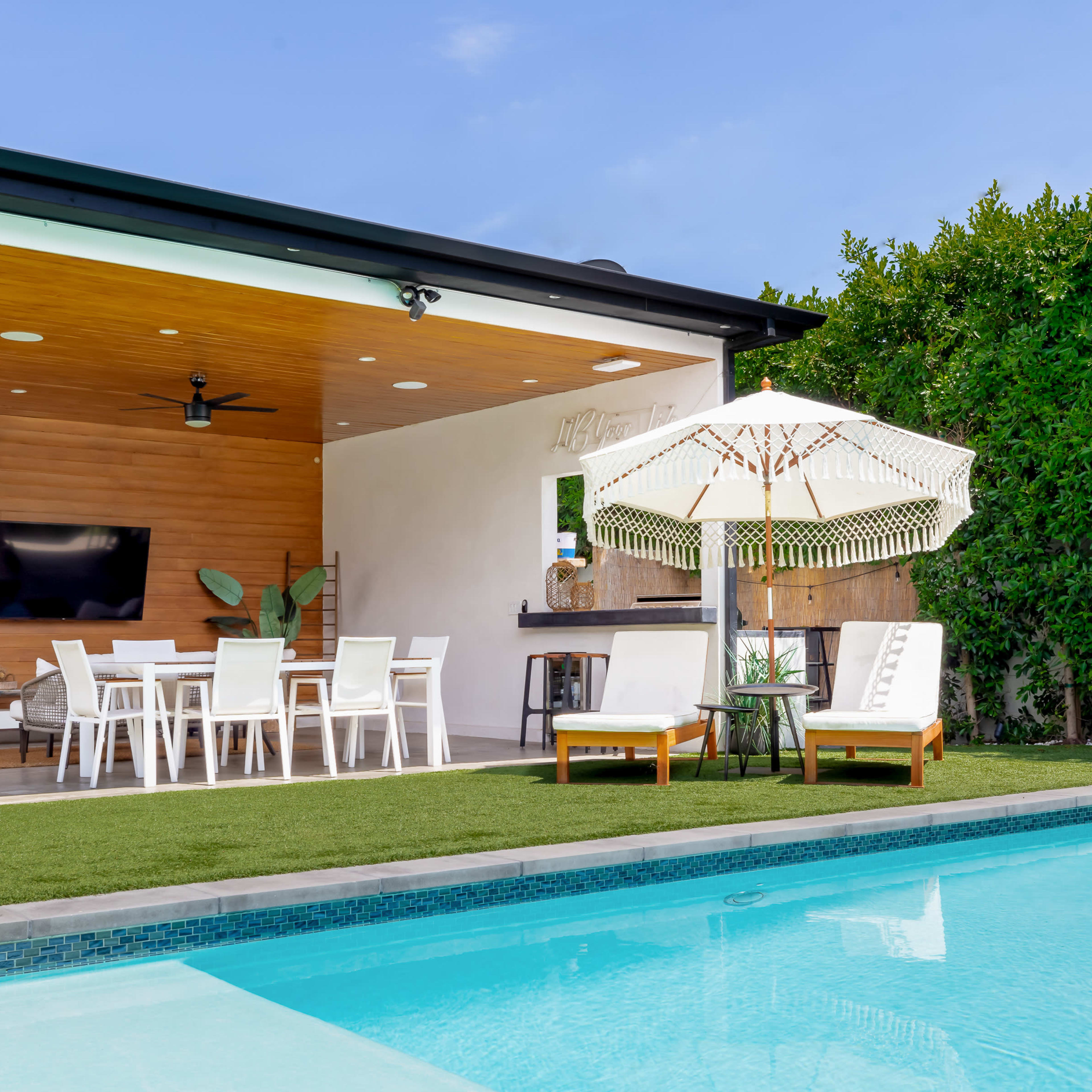 A modern outdoor living space with a covered patio, dining area, and a swimming pool surrounded by lush greenery.