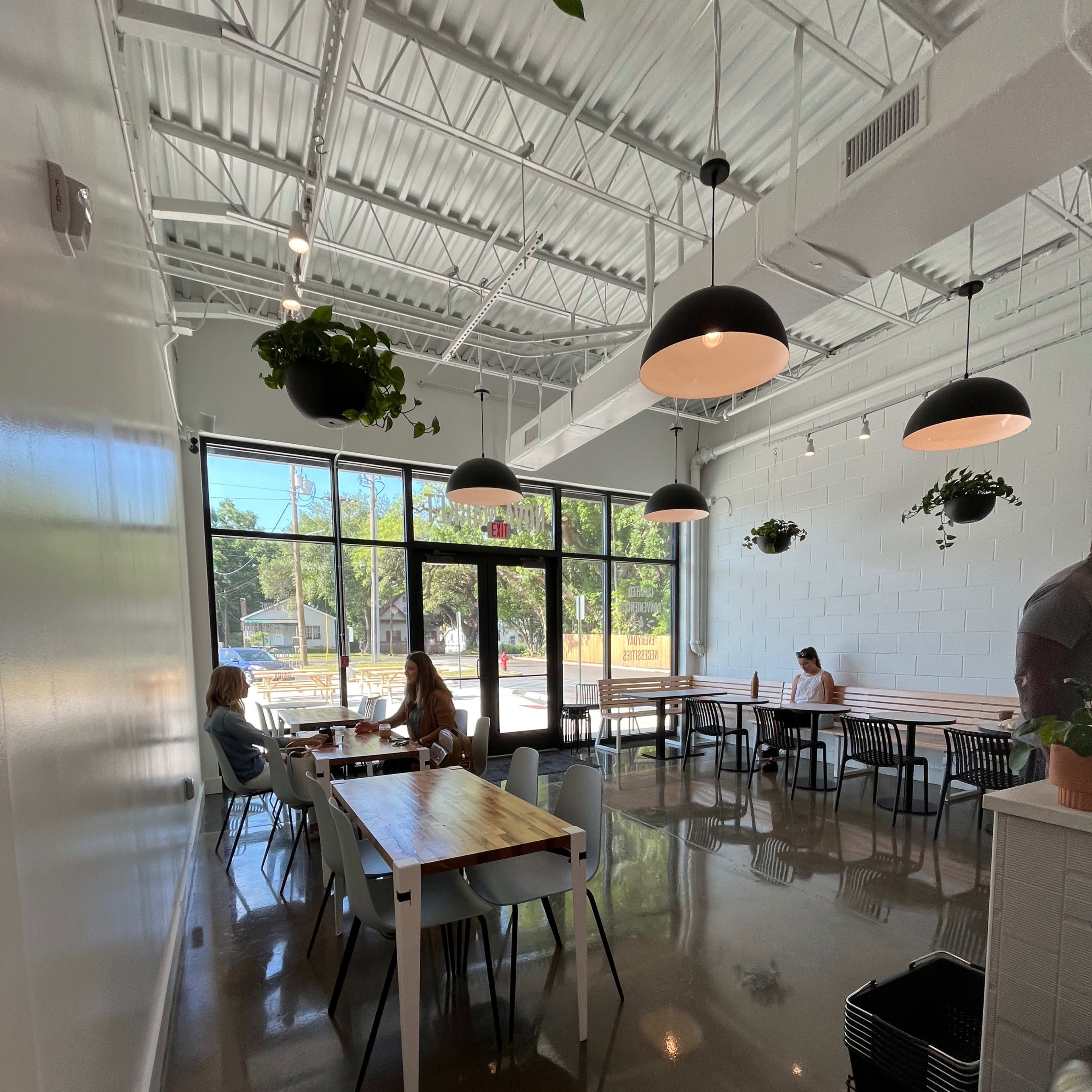 A bright, minimalist café with tables and chairs, featuring indoor plants and large windows overlooking the outside.