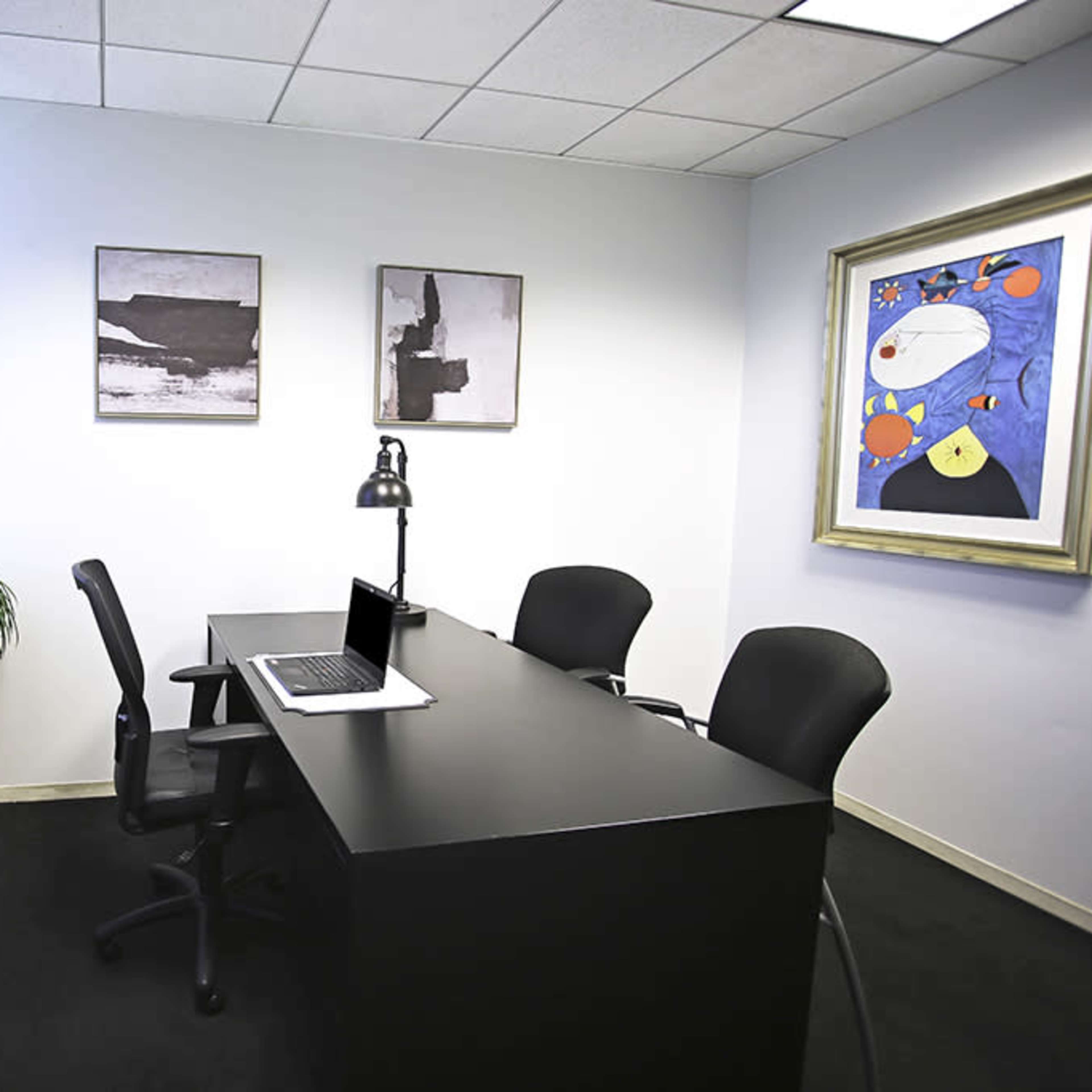 A modern office space with a black desk, two chairs, a laptop, and framed artworks on the walls.