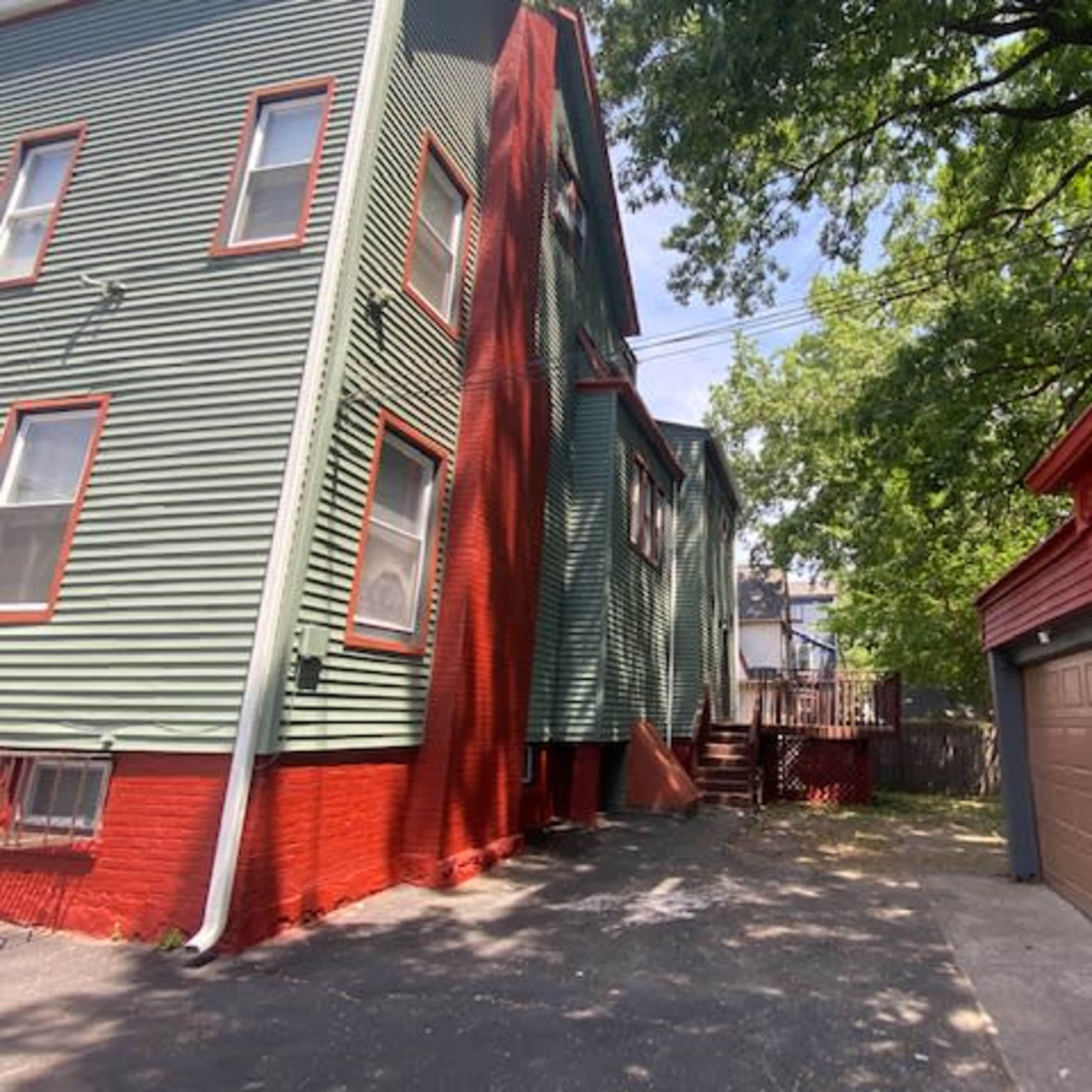 A narrow driveway between a green multi-story building and a red garage, with a deck visible at the back of the property.