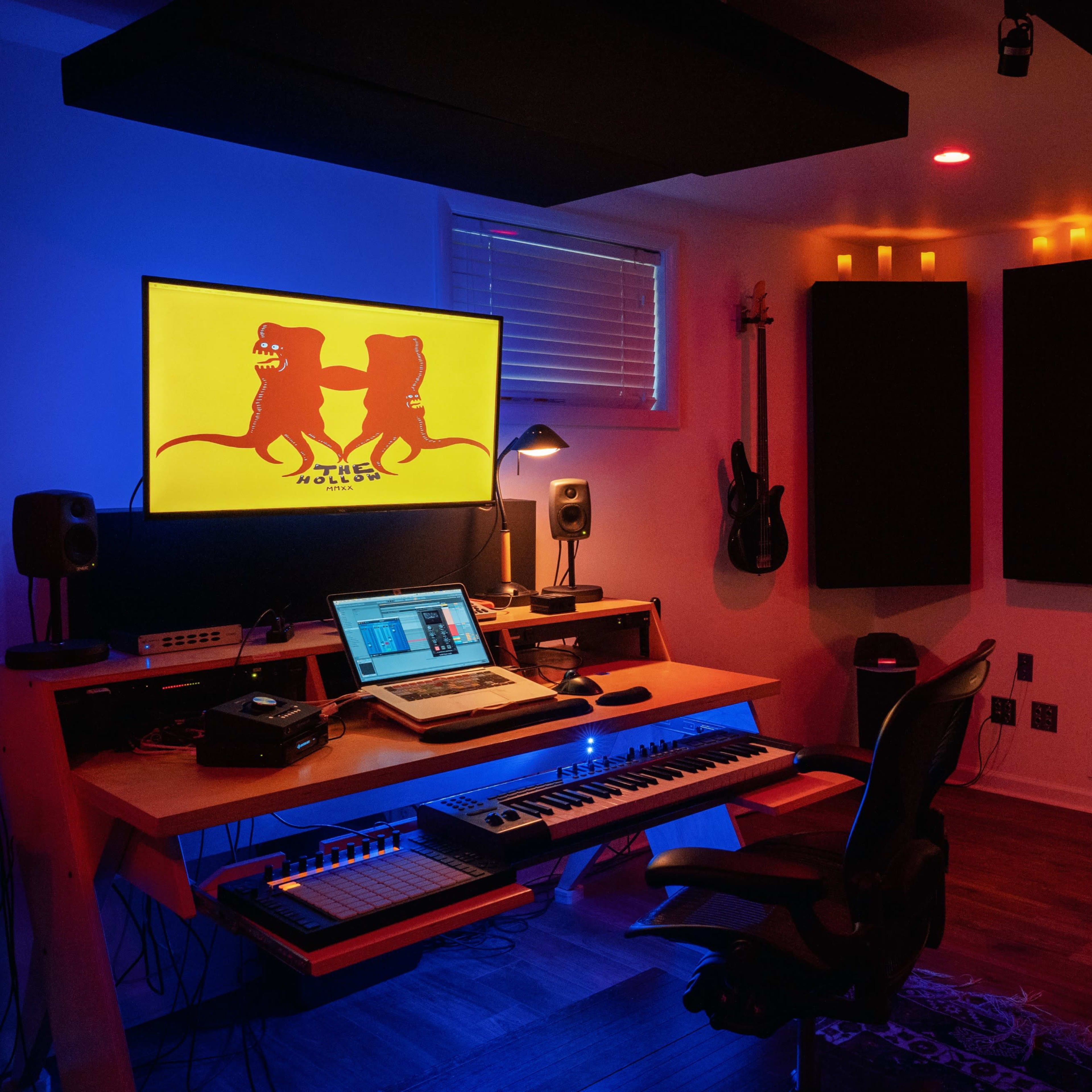 A dimly lit recording studio with a computer and MIDI keyboard on a wooden desk, surrounded by soundproof panels and colorful ambient lighting.