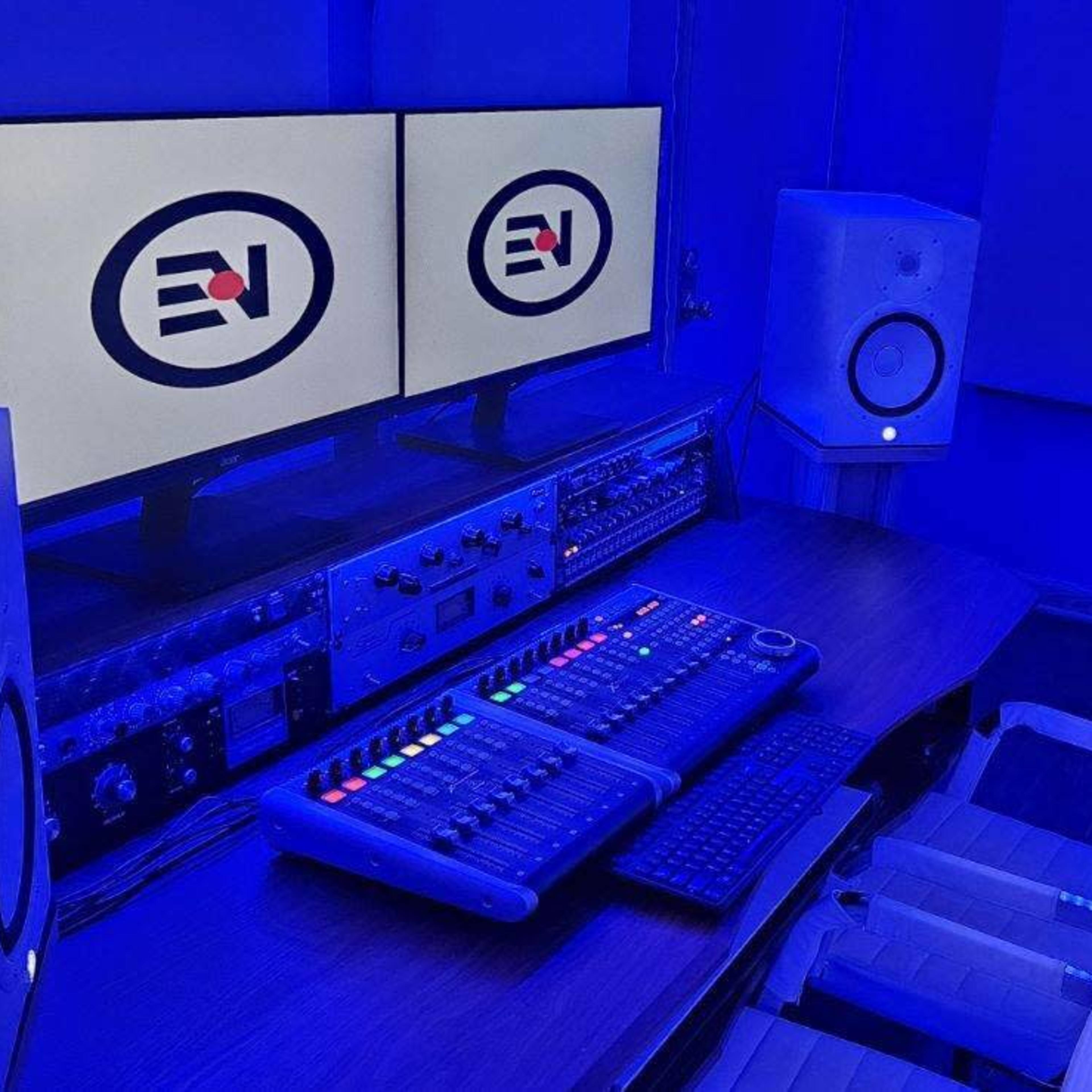 A music production studio features two monitors, a control console with colorful buttons, and studio monitors, all illuminated by blue lighting.